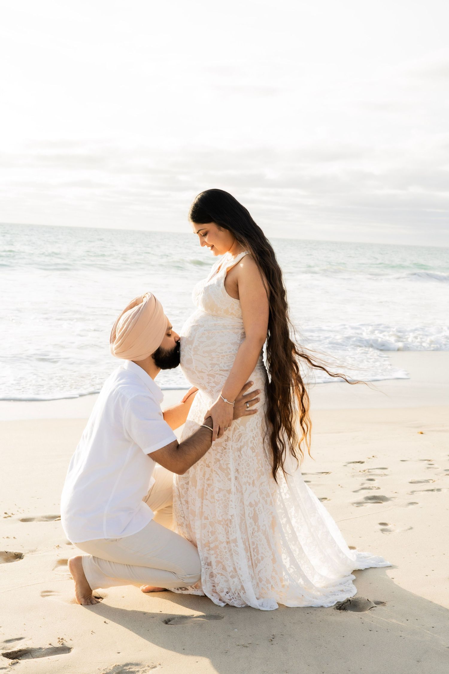 Man in turban kneeling on beach, lovingly holding pregnant woman's belly near ocean waves.