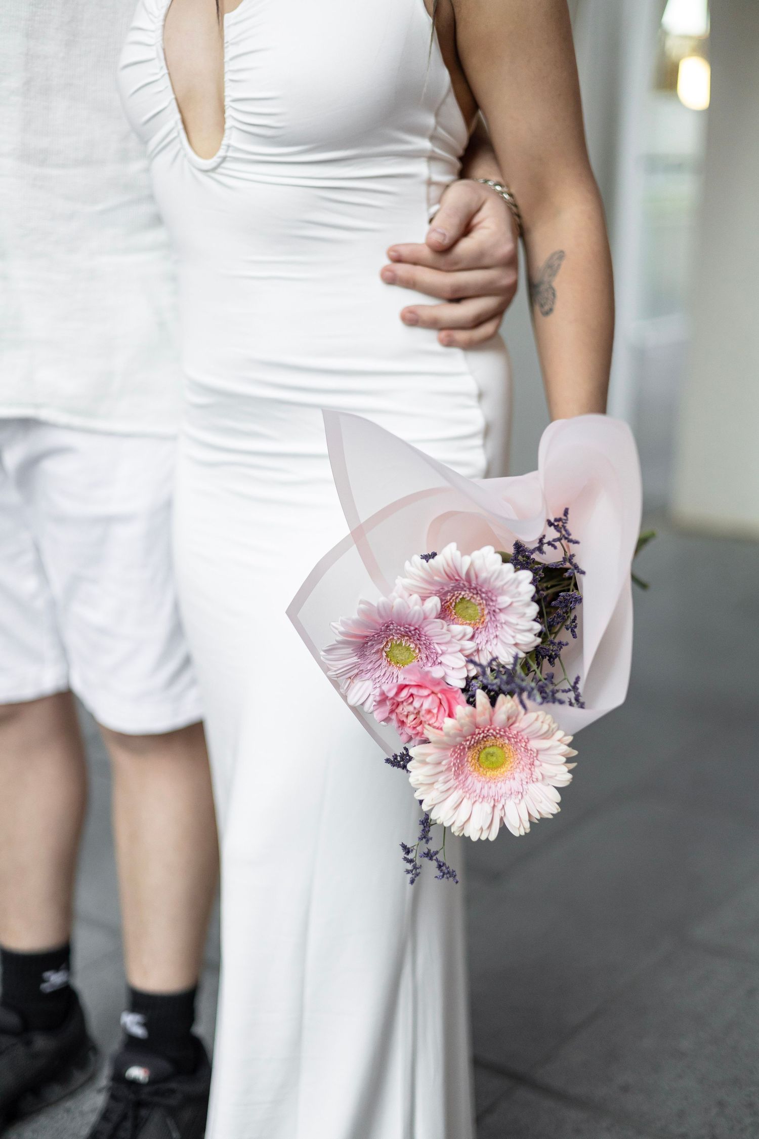 Pregnant woman in white halter dress holding pink gerbera daisy bouquet while partner embraces her waist.