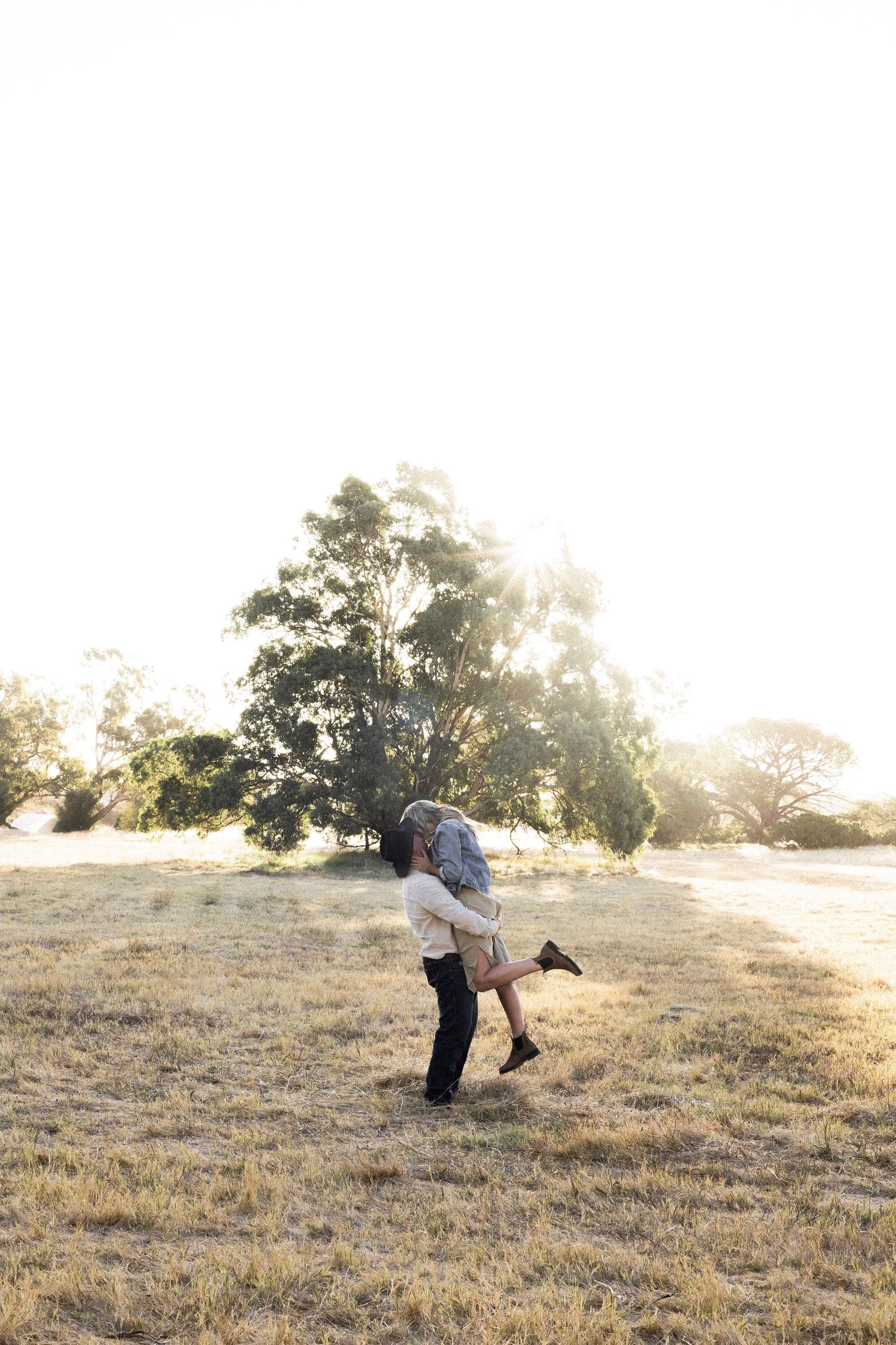 A couple shares a romantic lift and kiss in a golden sunlit field with trees glowing in the background.
