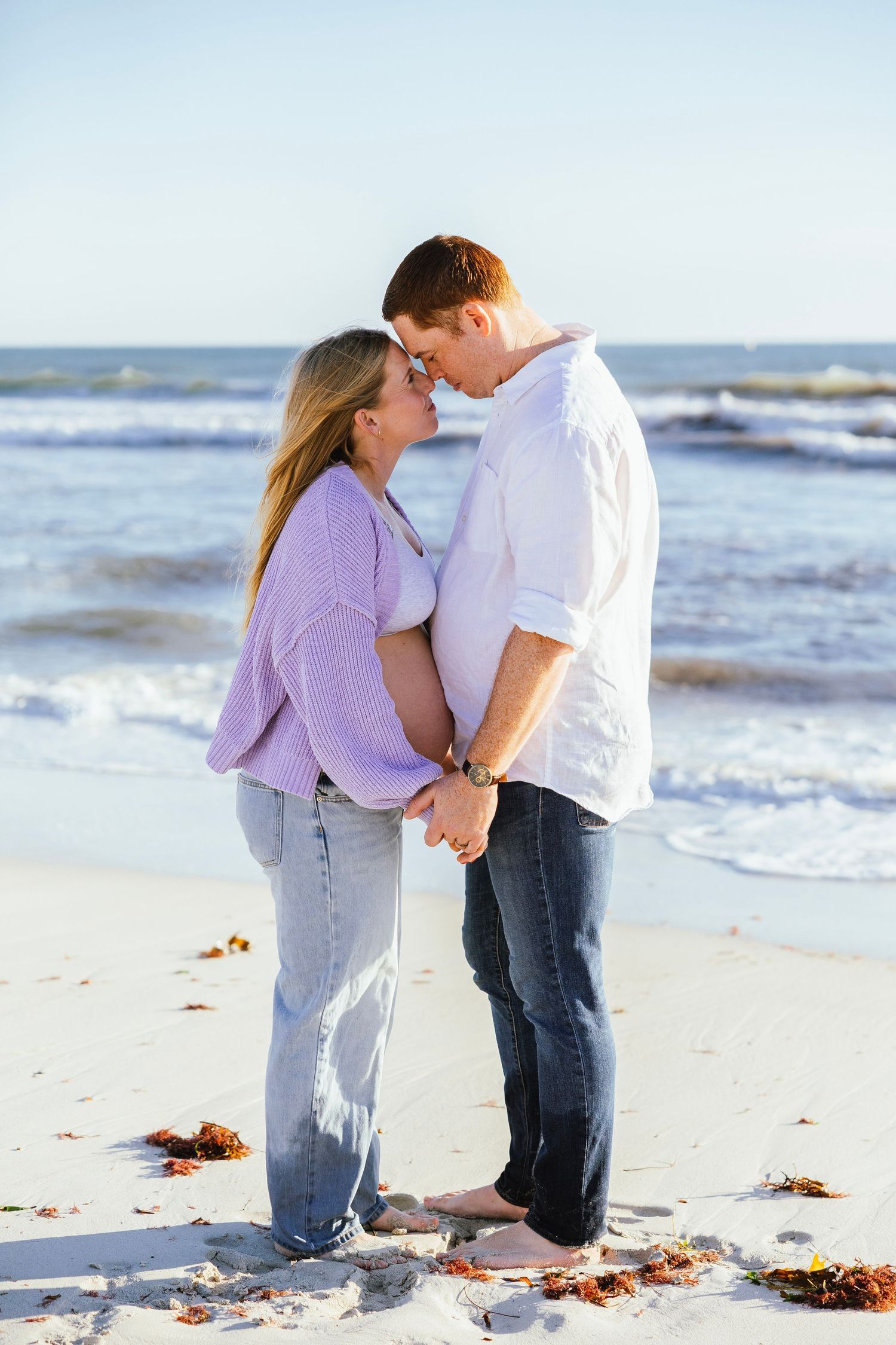 Expecting couple holding hands and touching foreheads tenderly on sandy beach with ocean waves behind.