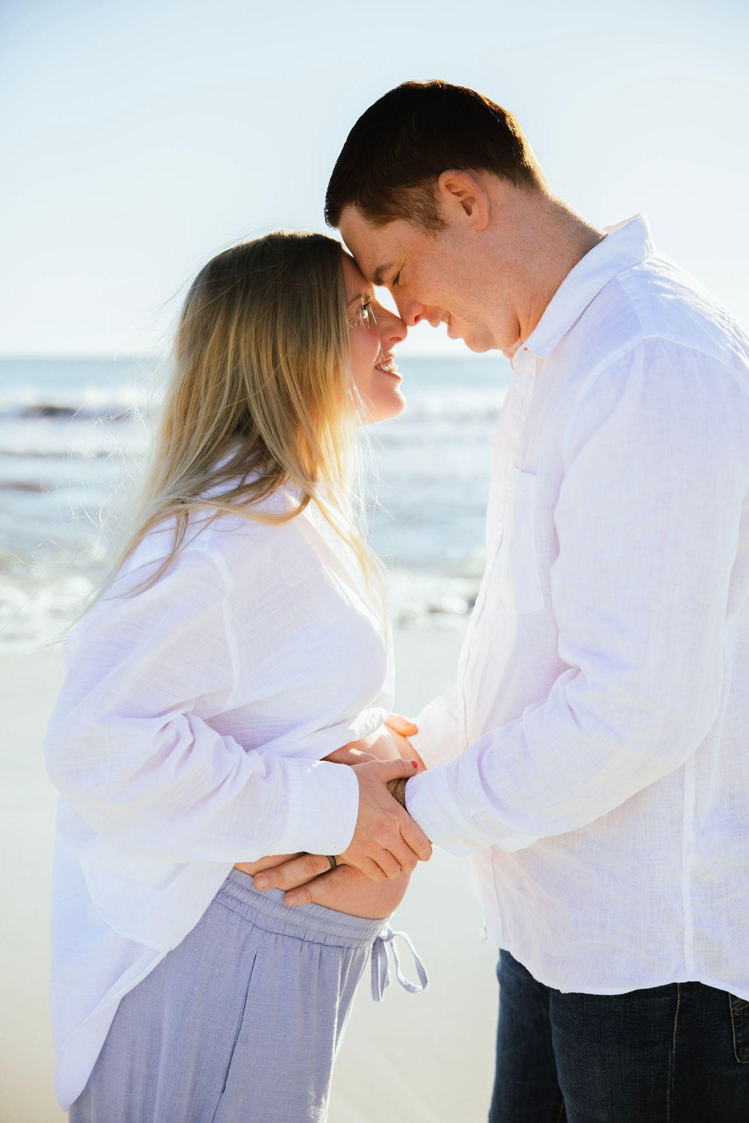Pregnant couple in white shirts tenderly touching foreheads on a sunny beach, holding hands over baby bump.