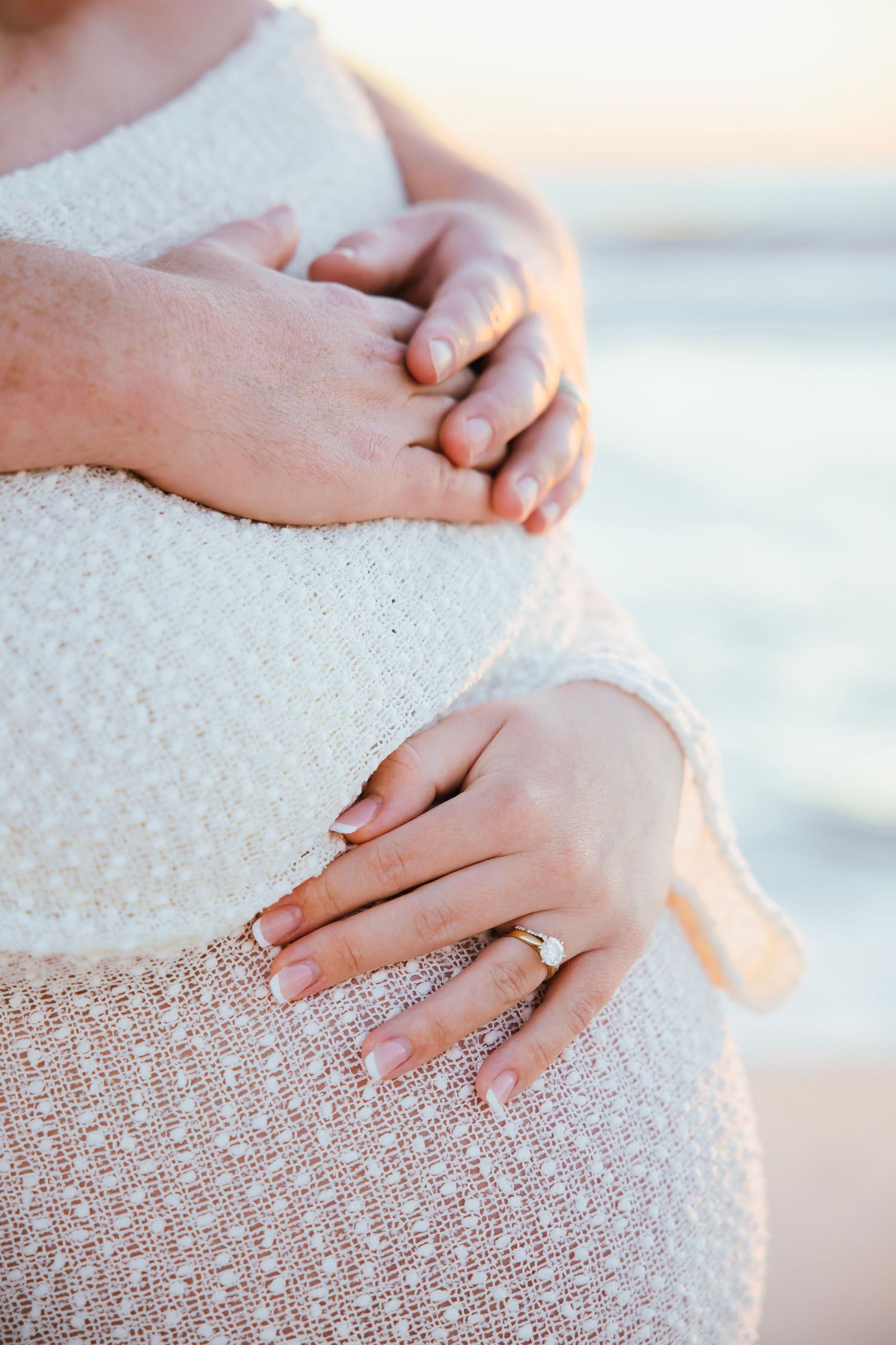 Close-up of couple's hands tenderly holding pregnant belly in white knit dress with engagement ring.