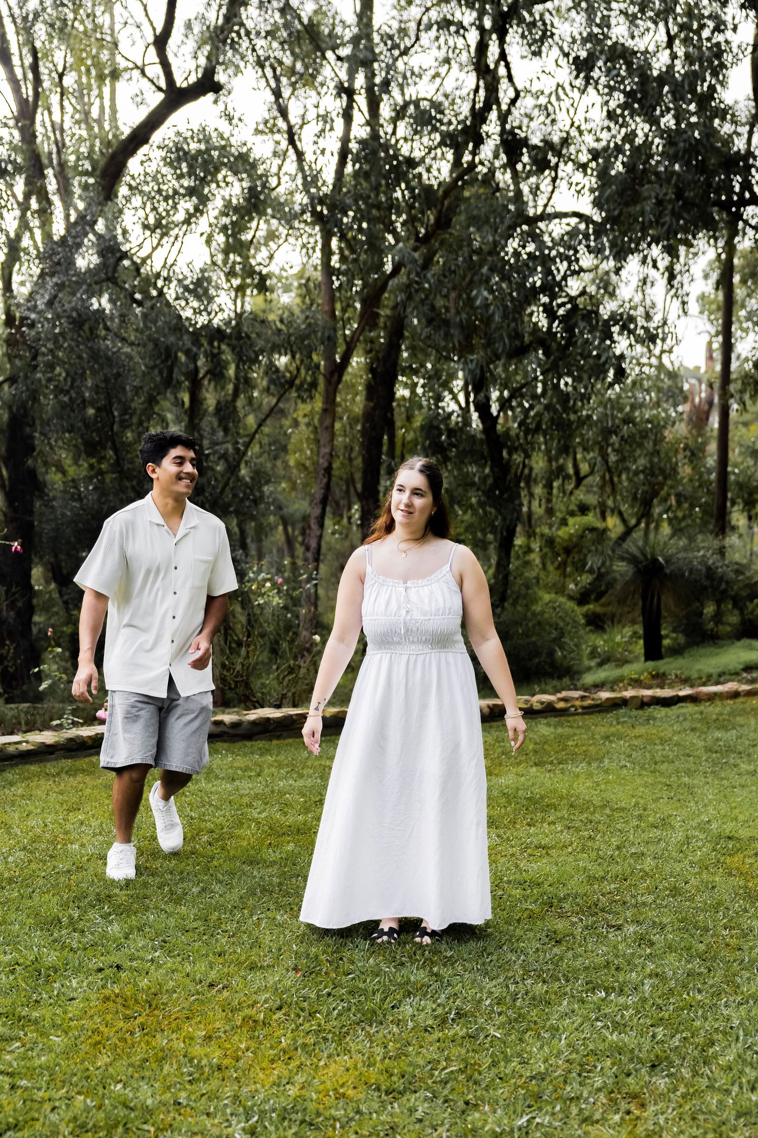A couple walks hand in hand across a green lawn surrounded by tall trees in a scenic outdoor setting.