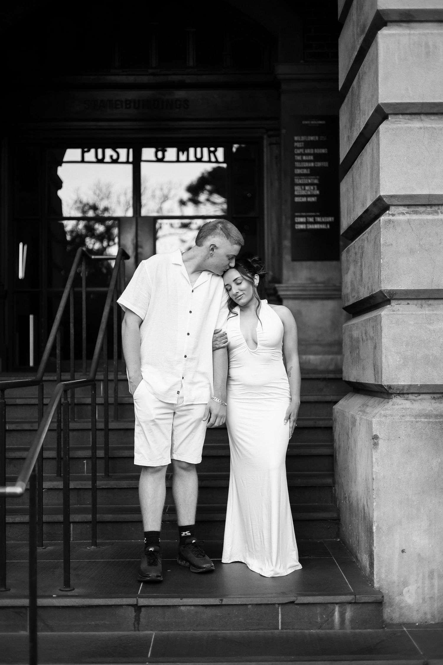 Black and white photo of a couple in white outfits embracing on steps outside a historic building.
