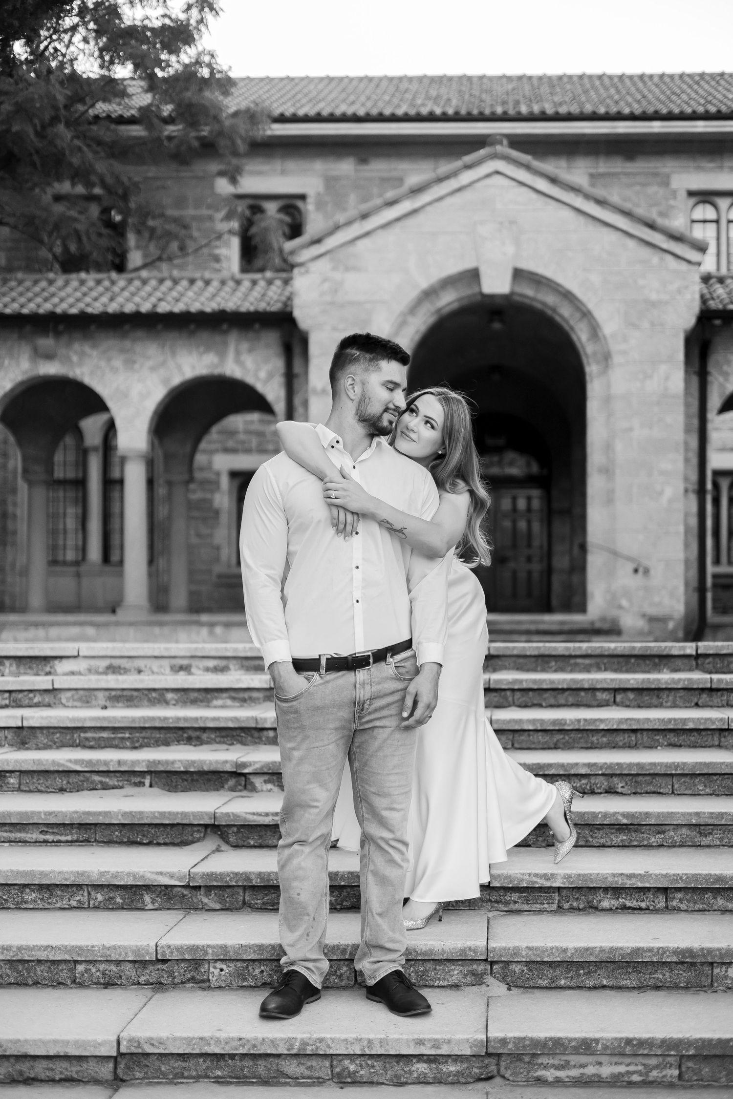 Black and white photo of a couple embracing on stone steps outside a historic arched stone building.