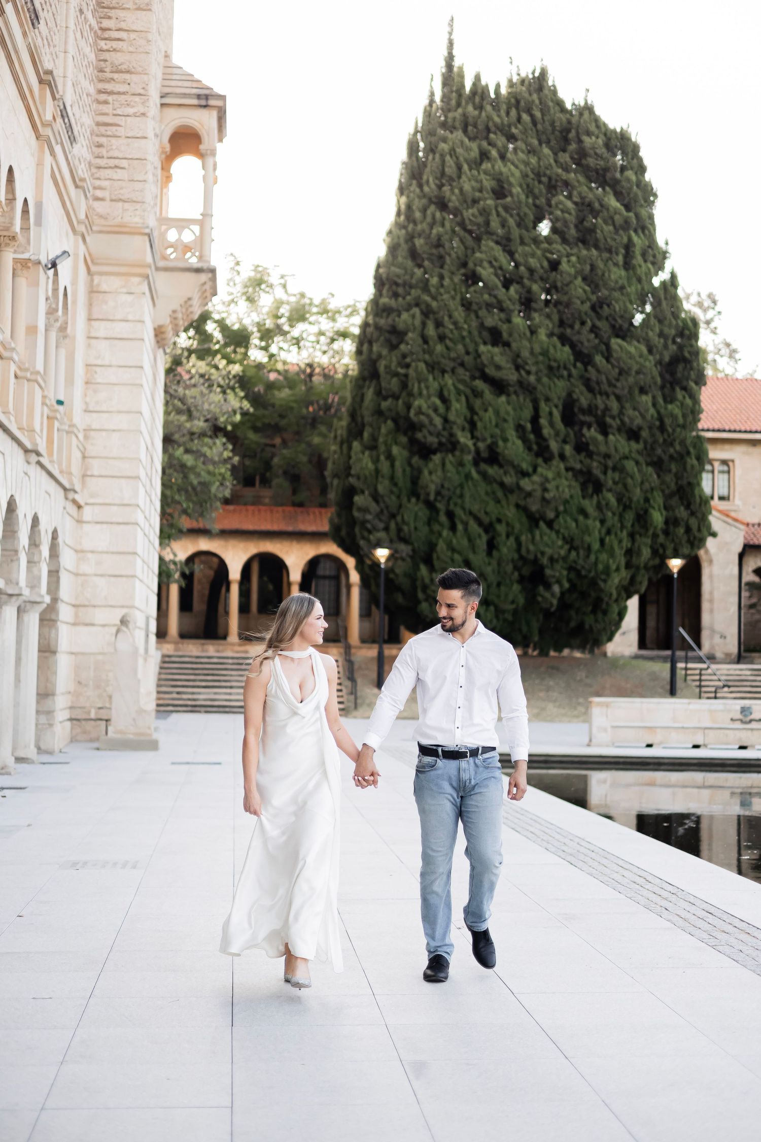 Couple holding hands walking along elegant stone courtyard with arched building and cypress tree backdrop.