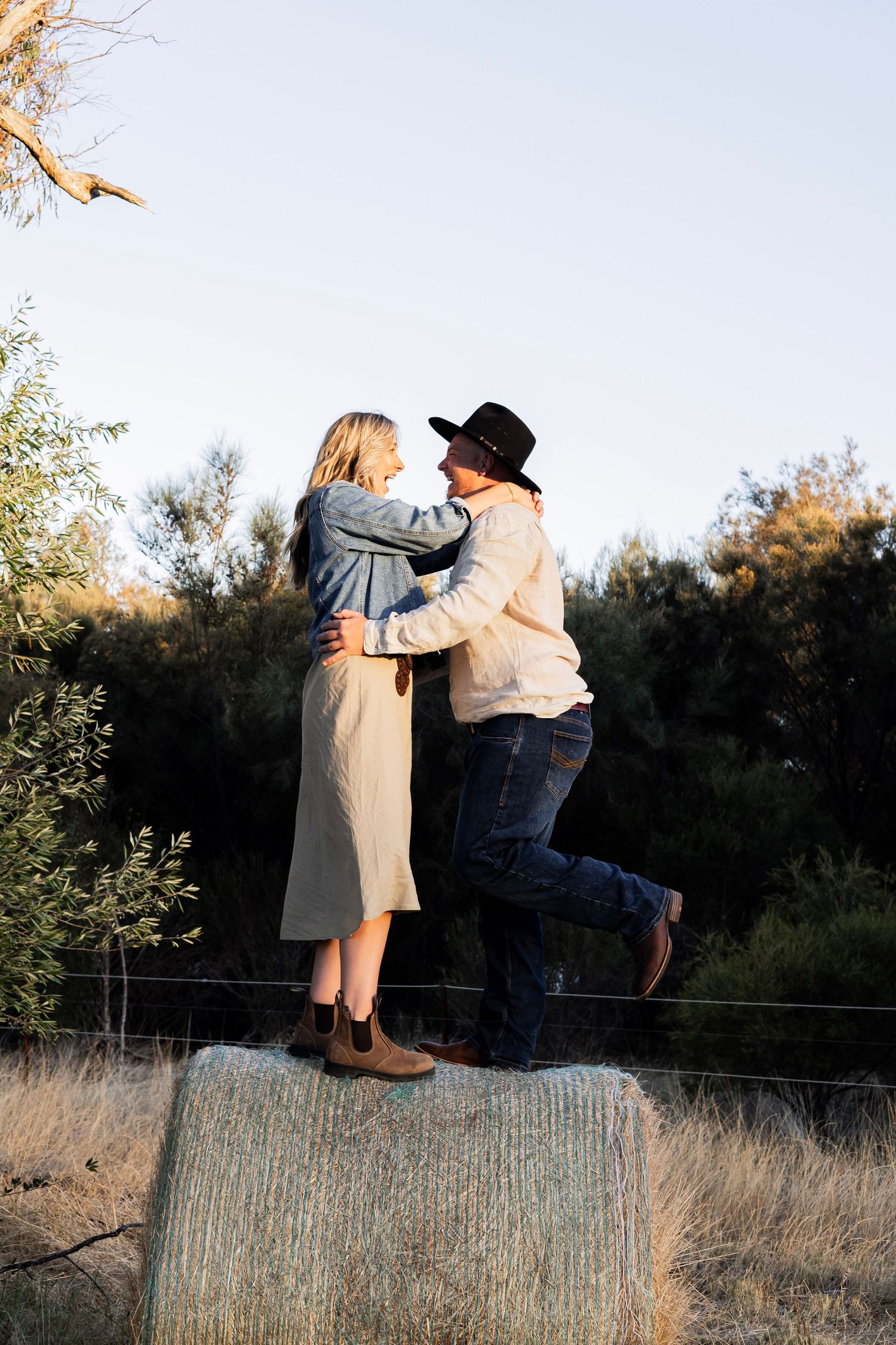 A couple embraces on a hay bale at golden hour, surrounded by trees and dry grass.