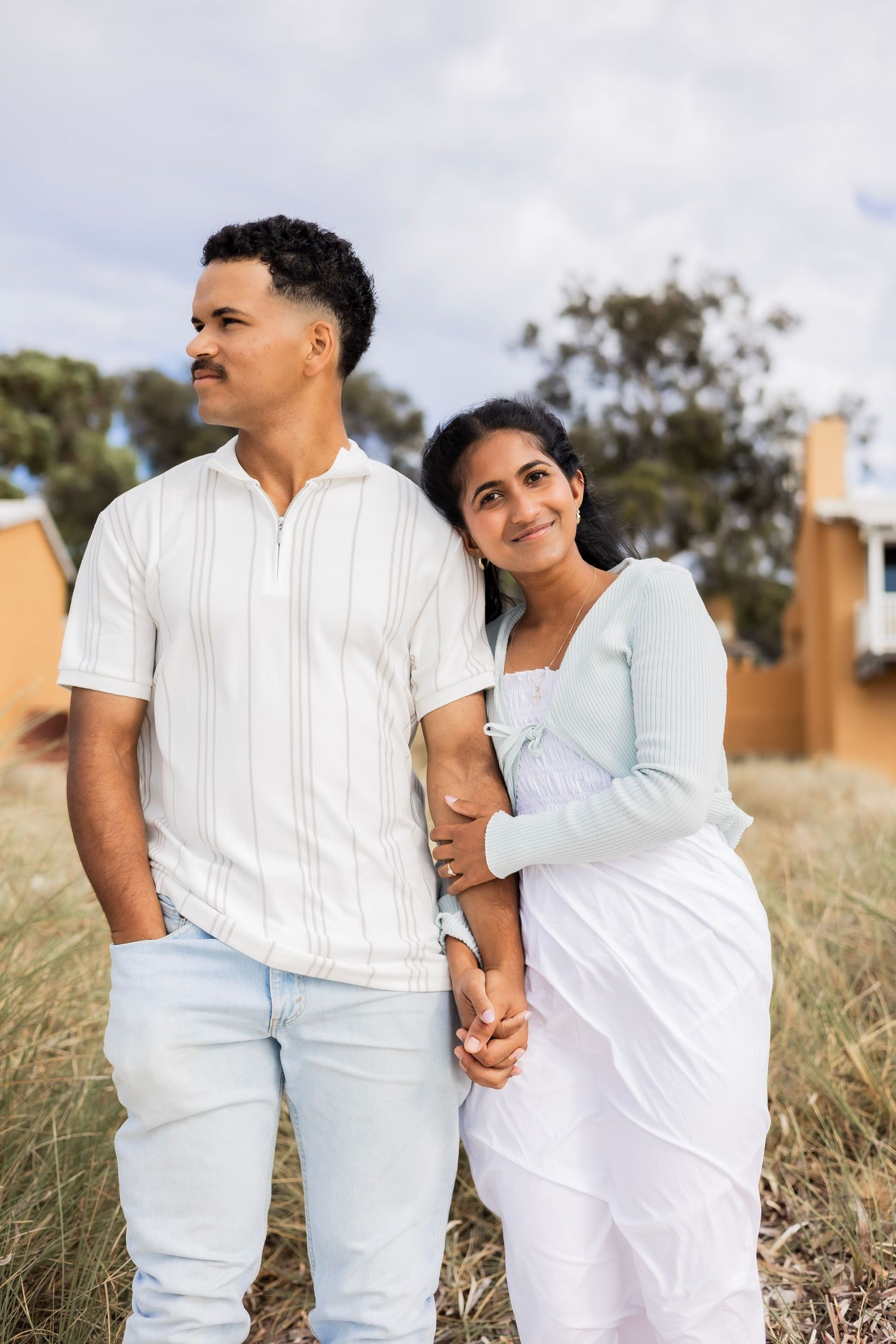 Couple standing in coastal field, woman leaning on mans arm, both looking into the distance.