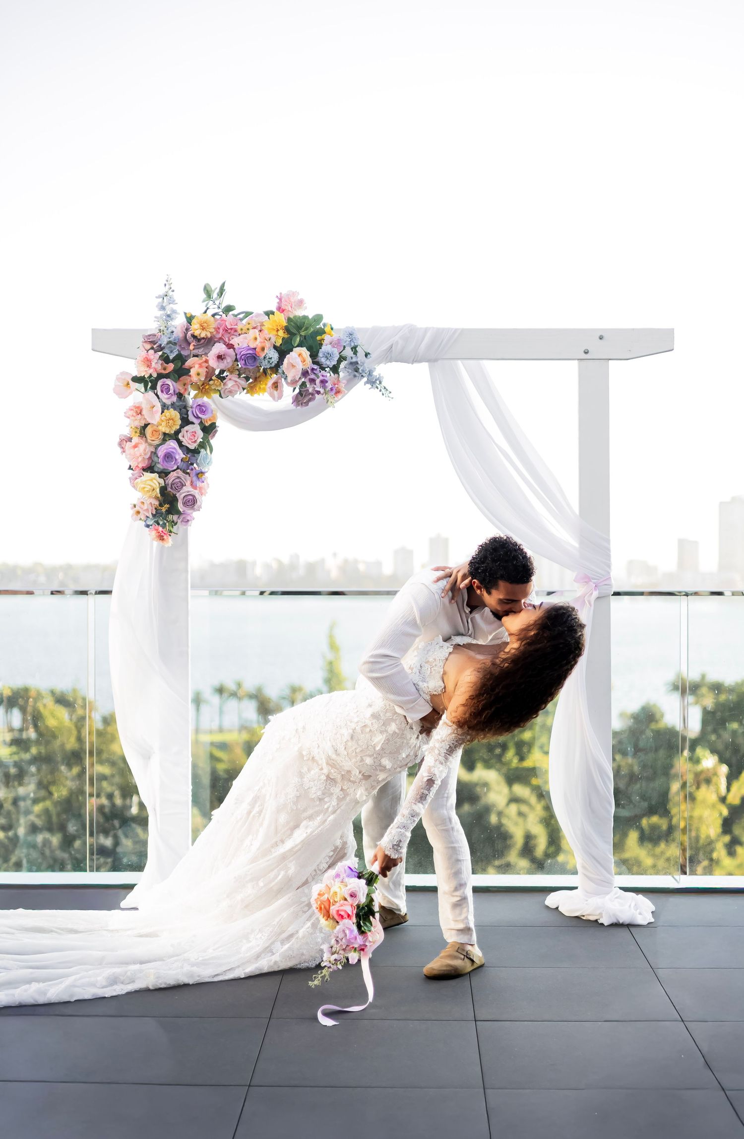 Bride and groom share dip kiss under colorful floral wedding arch on rooftop venue.