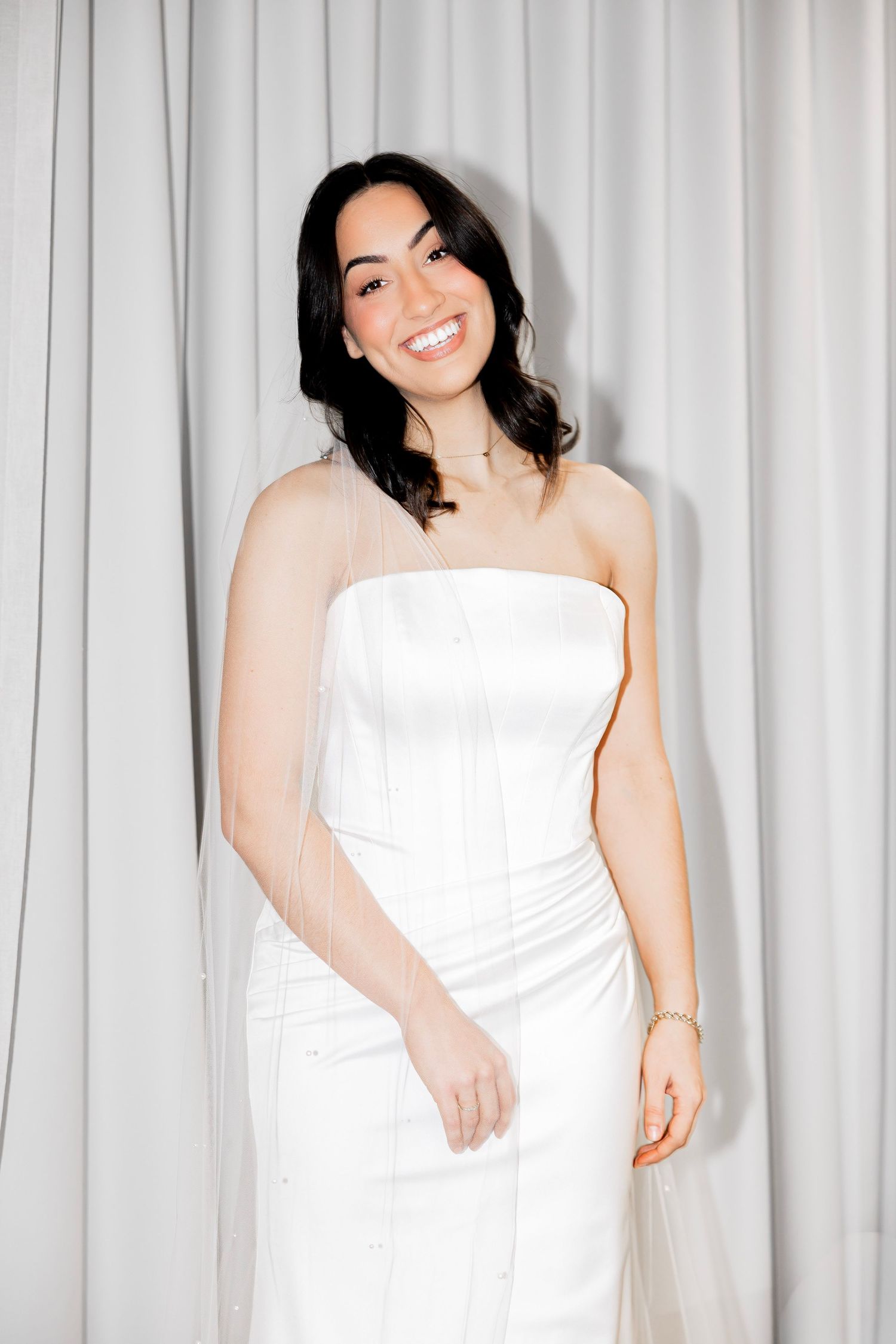 Smiling young woman in a white strapless corset dress posing in front of white curtains.