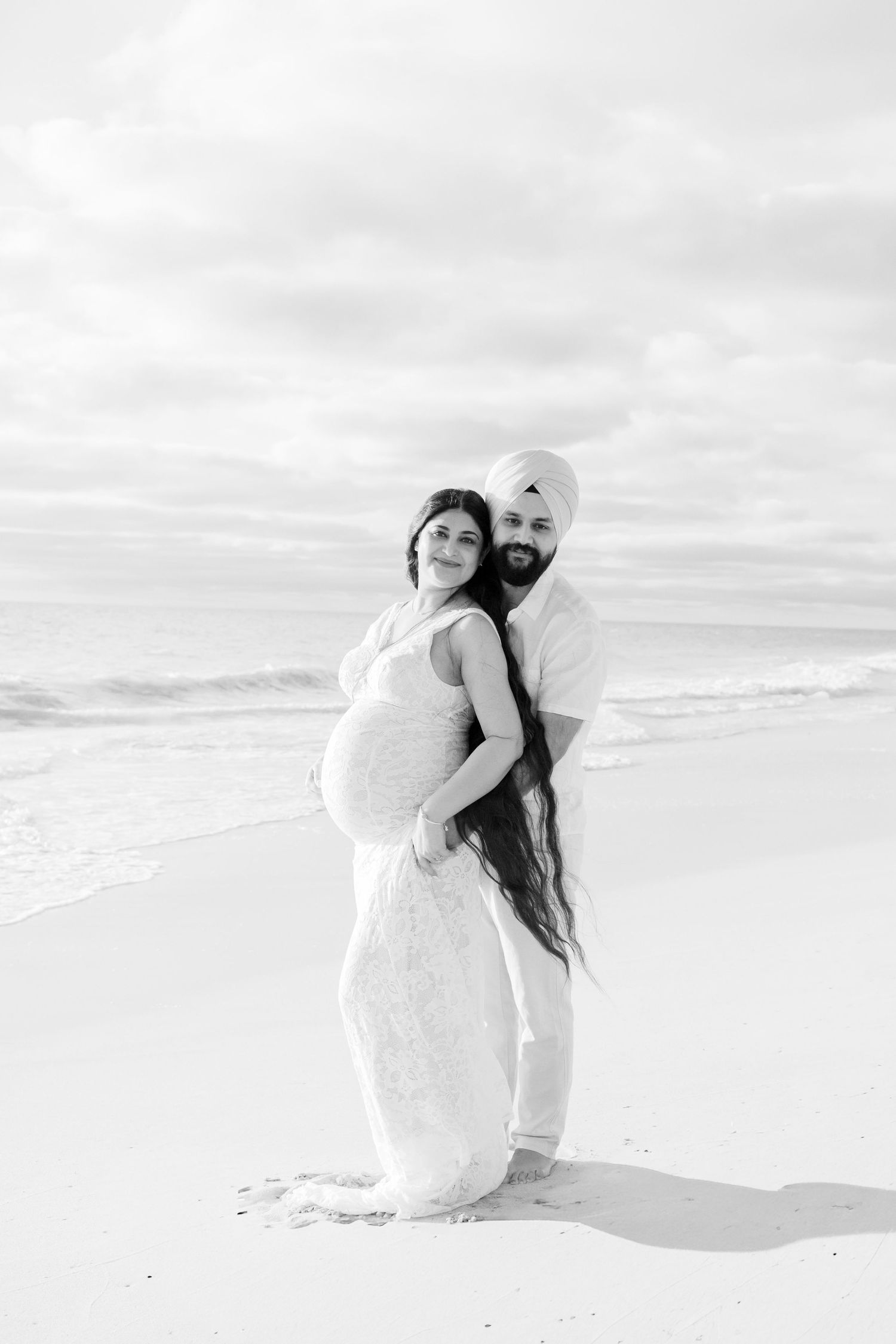 Black and white maternity photo of couple on beach, man in turban hugging pregnant woman from behind.