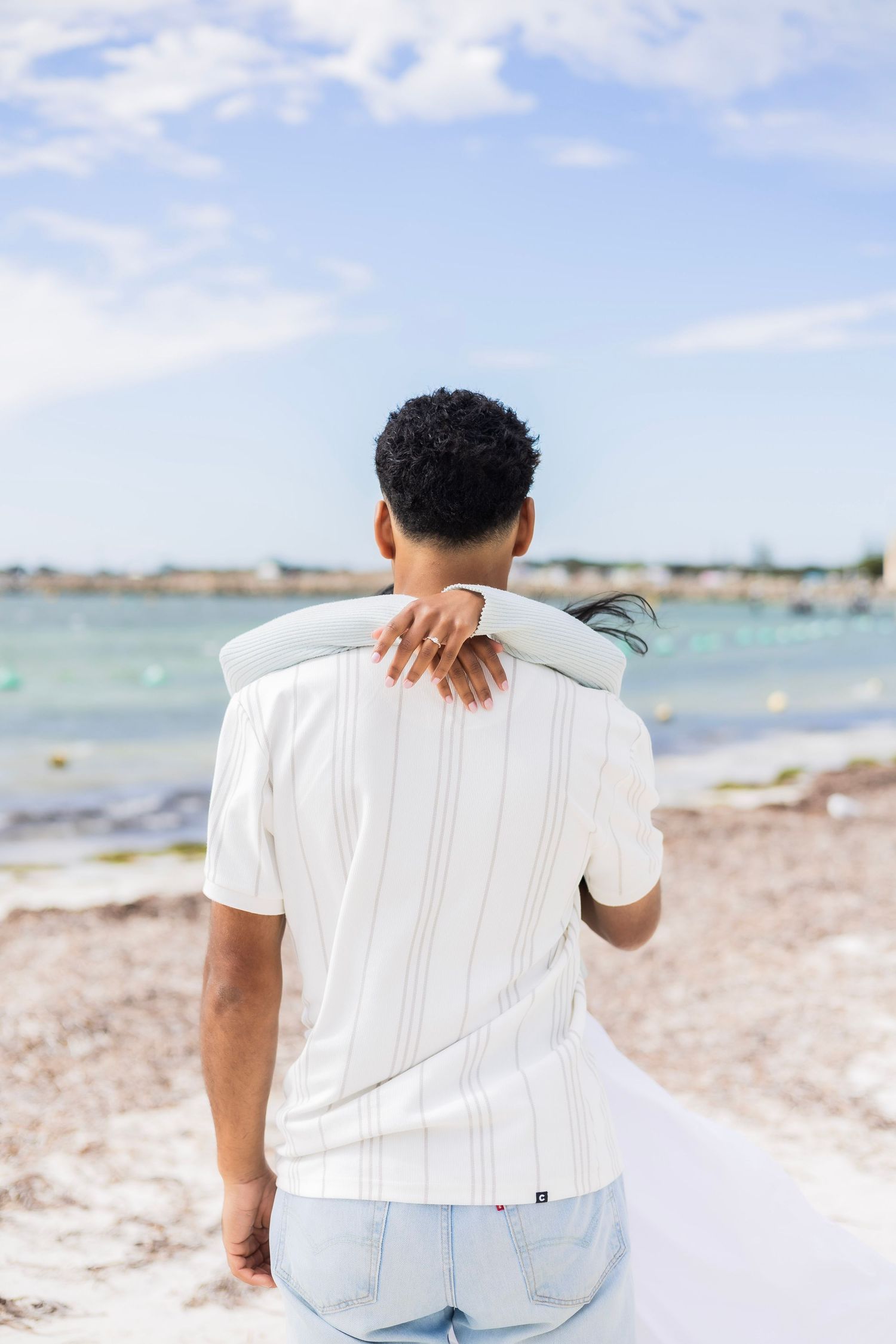 A man in a white shirt stands at the beach as a woman rests her arm on his shoulder.