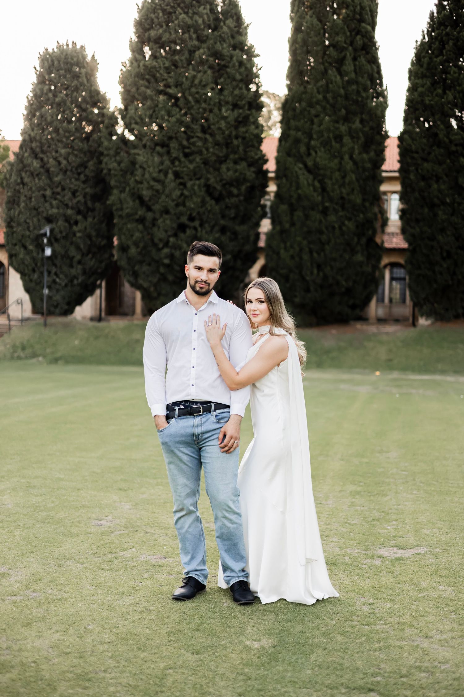 Engaged couple posing on green lawn with tall cypress trees, woman in white gown touching mans chest.