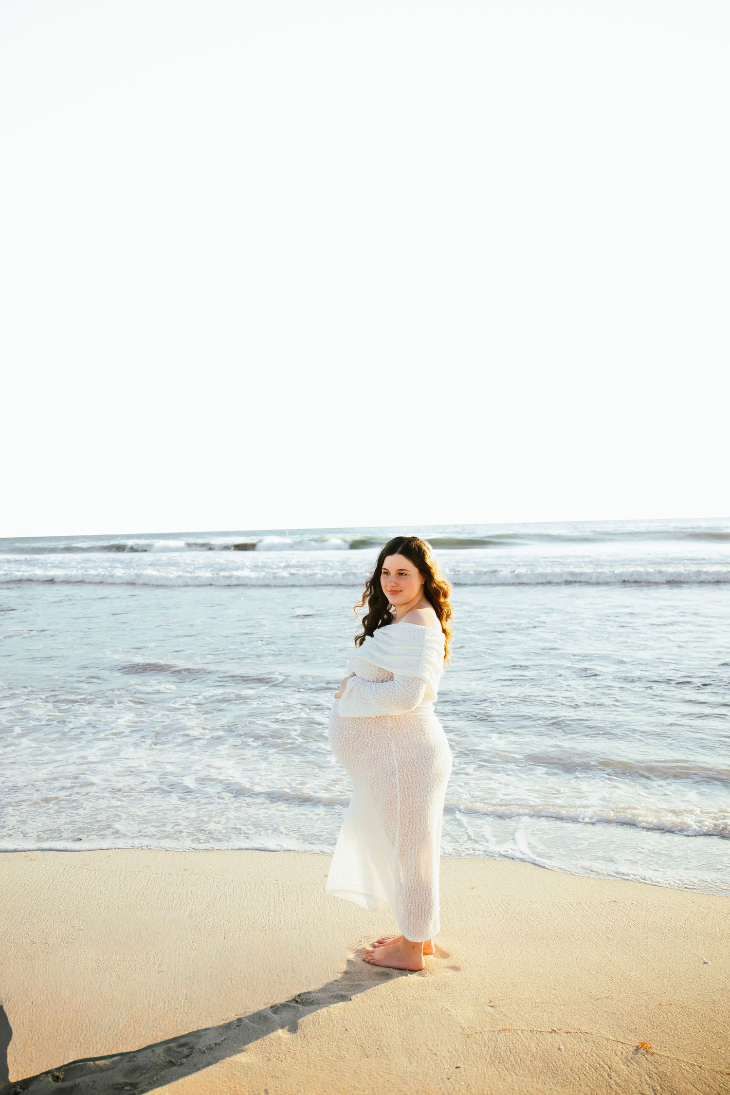 A woman in a flowing white dress stands barefoot on a sandy beach with ocean waves behind her.
