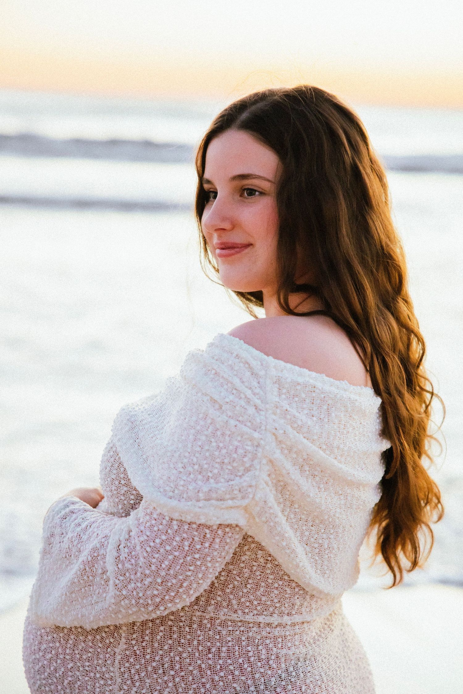 Pregnant woman in off-shoulder white knit dress posing on beach at golden hour sunset.