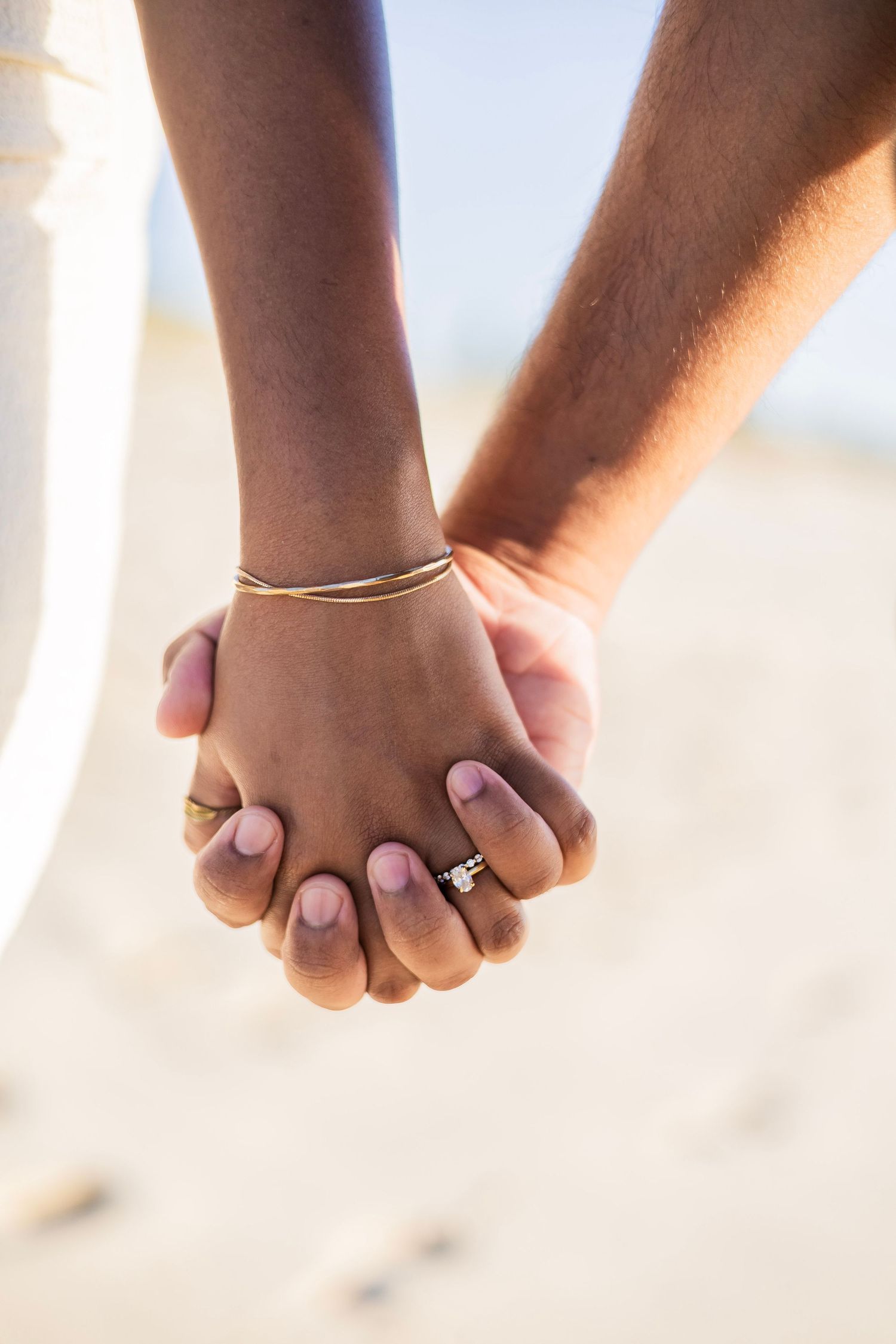 Close-up of an interracial couple holding hands on a beach, woman wearing an engagement ring and gold bracelet.
