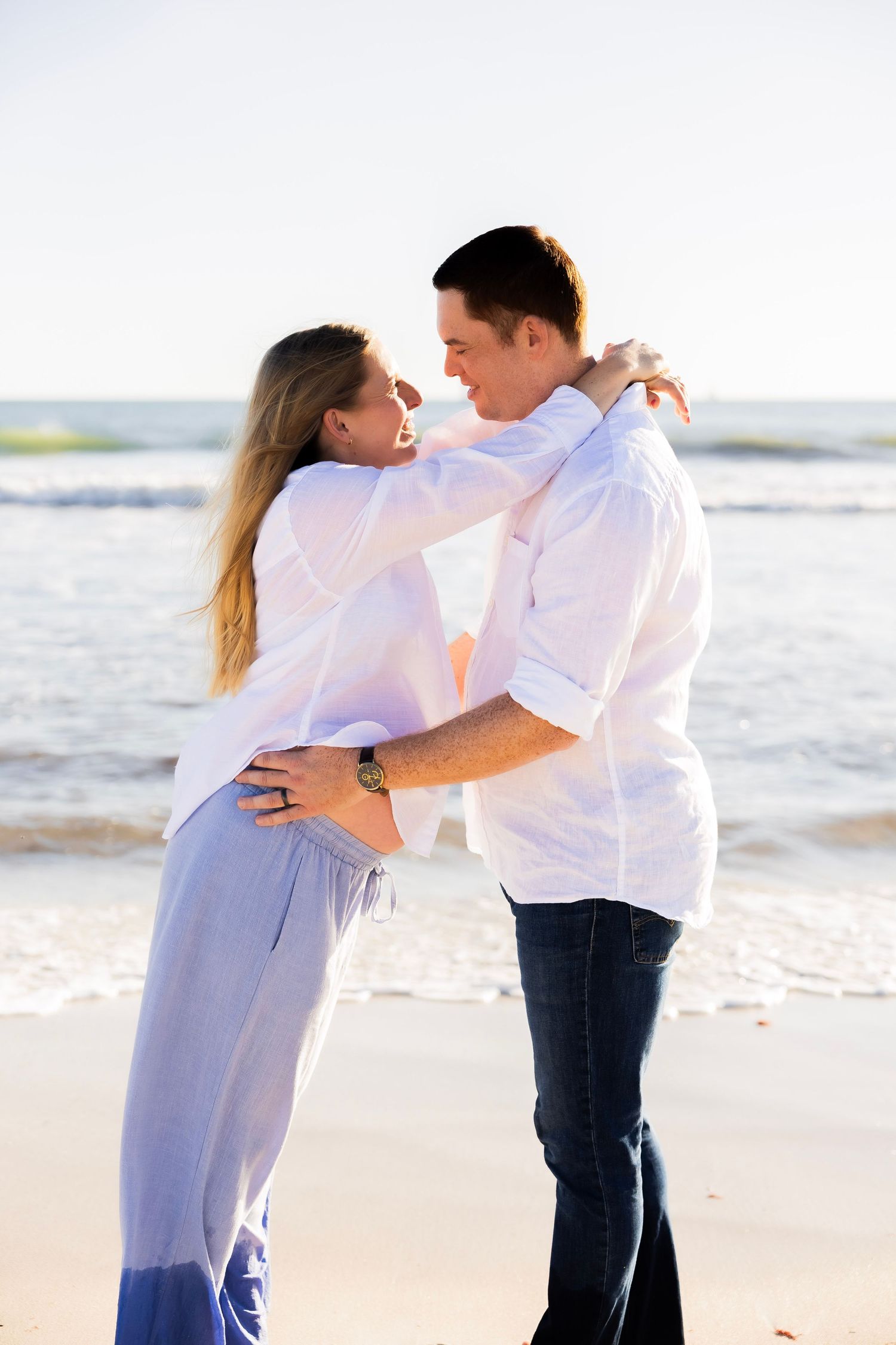 Couple embracing and gazing at each other romantically on a sunny beach with ocean waves behind them.