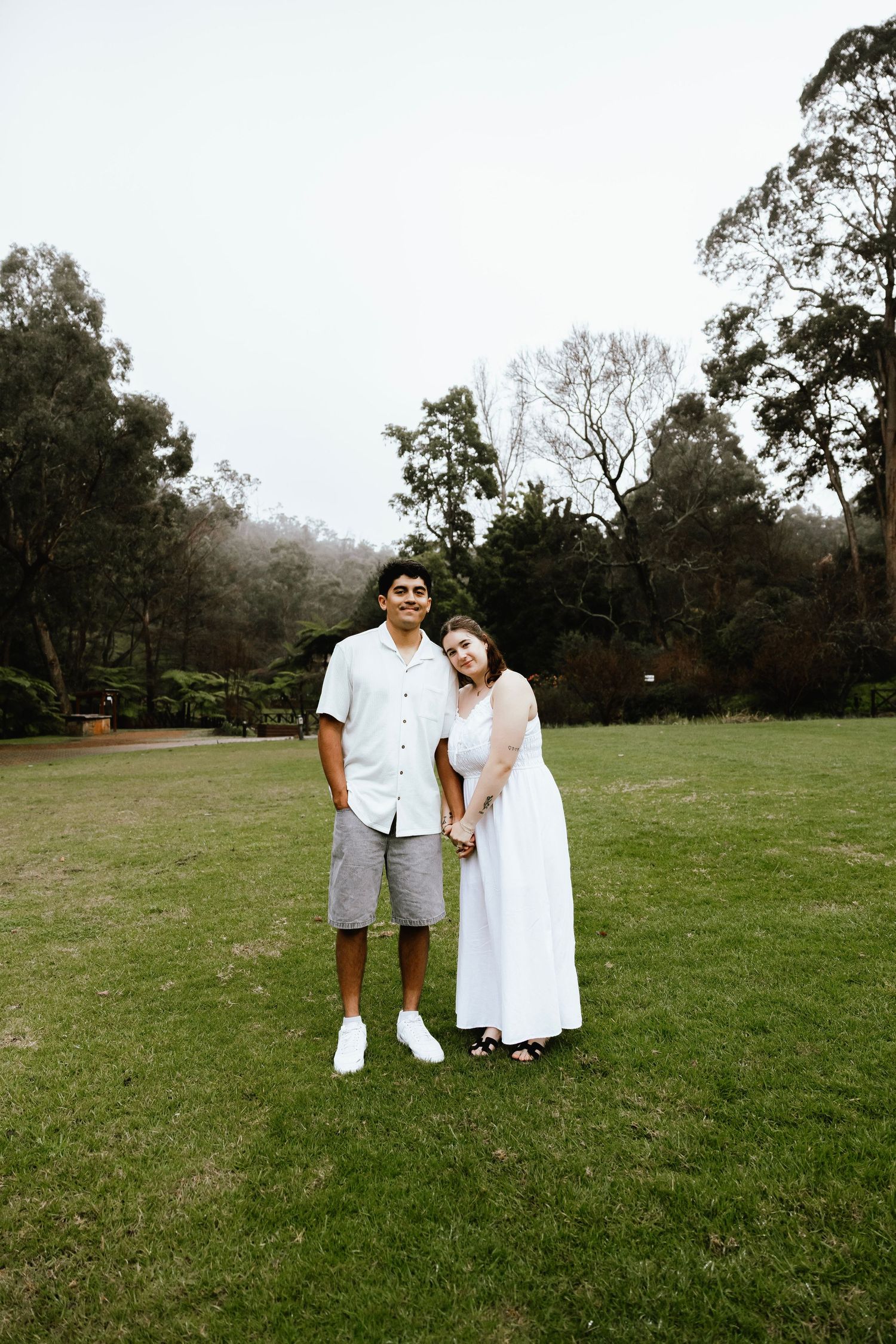 Smiling couple standing together on green grass with misty trees in the background.