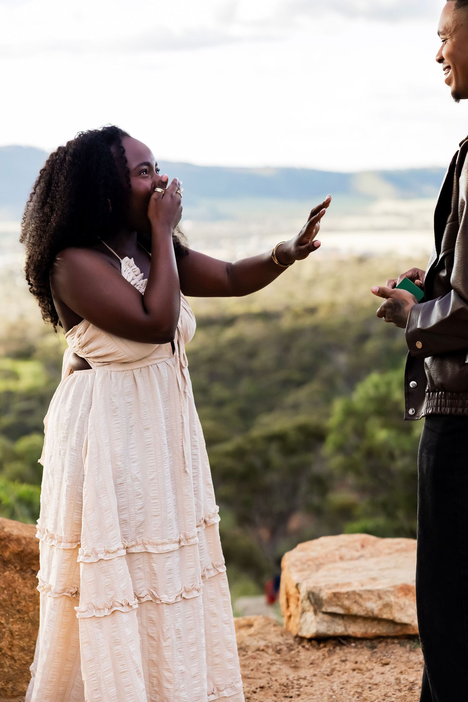 A surprised woman in a white dress covers her mouth during an outdoor marriage proposal with scenic landscape views.