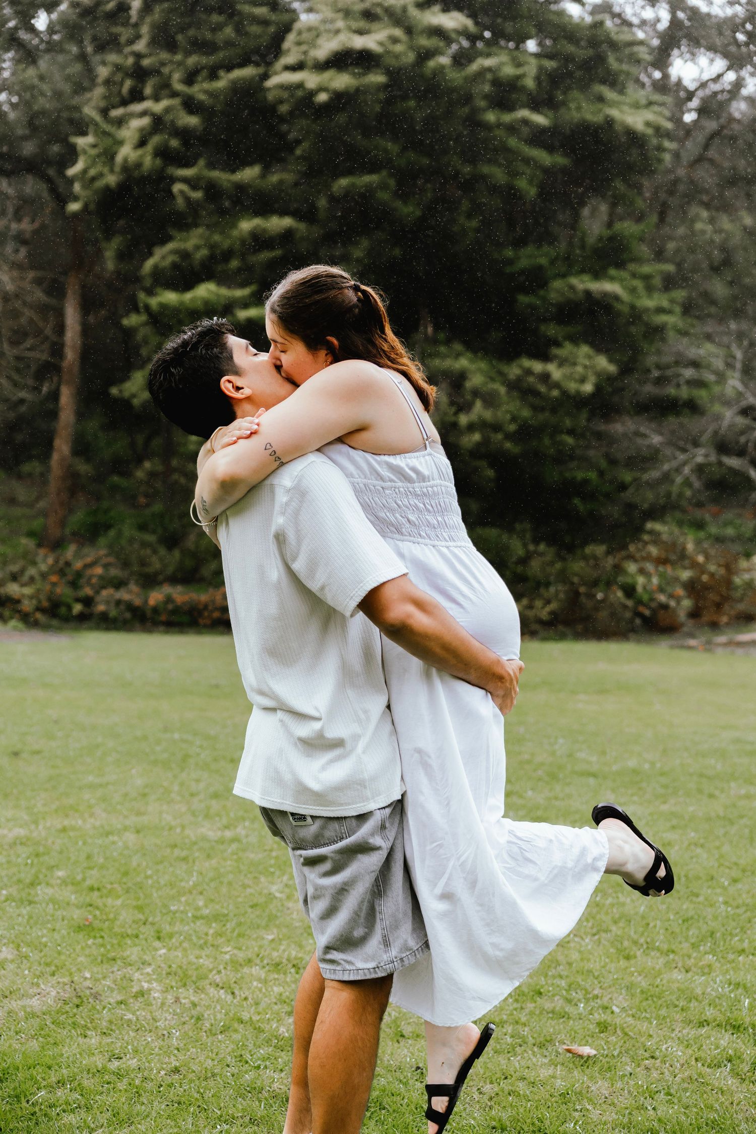 A couple shares a romantic kiss in a lush green park, as the man lifts the woman in a white dress off the ground.