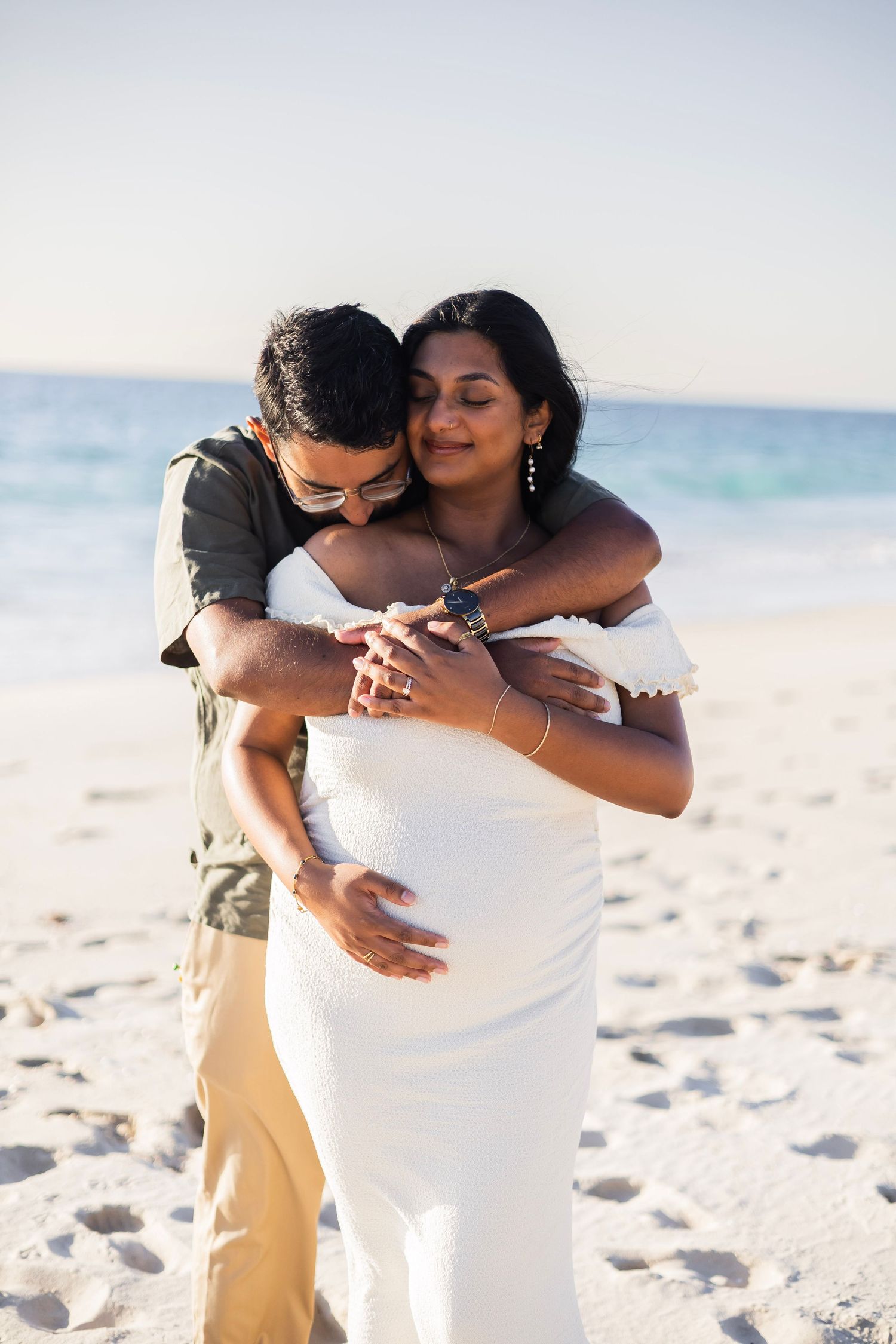 Man embracing pregnant woman from behind on a sandy beach, both smiling warmly in golden sunlight.