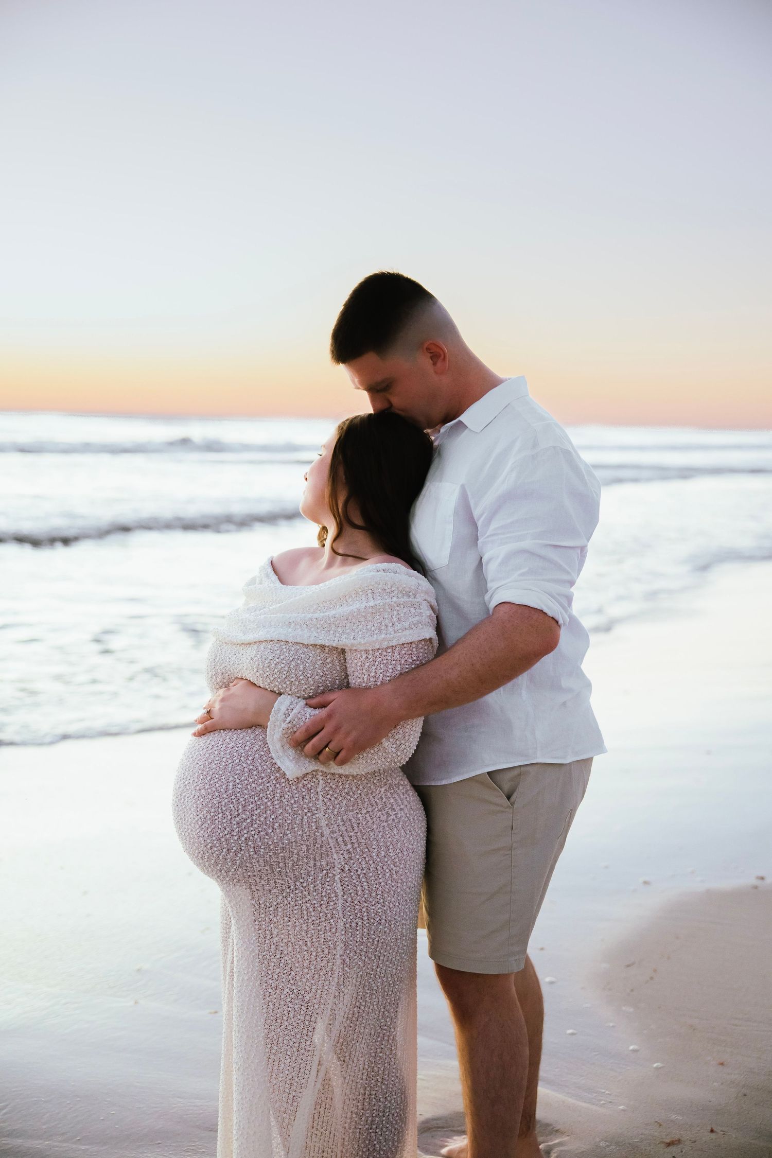 Couple sharing tender moment on beach at sunset, man embracing pregnant partner in white lace dress.