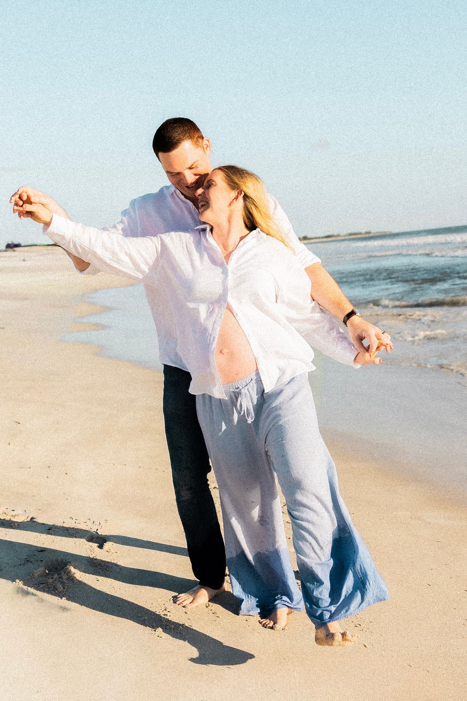 Happy pregnant couple dancing barefoot on sandy beach in white shirts near ocean shoreline.