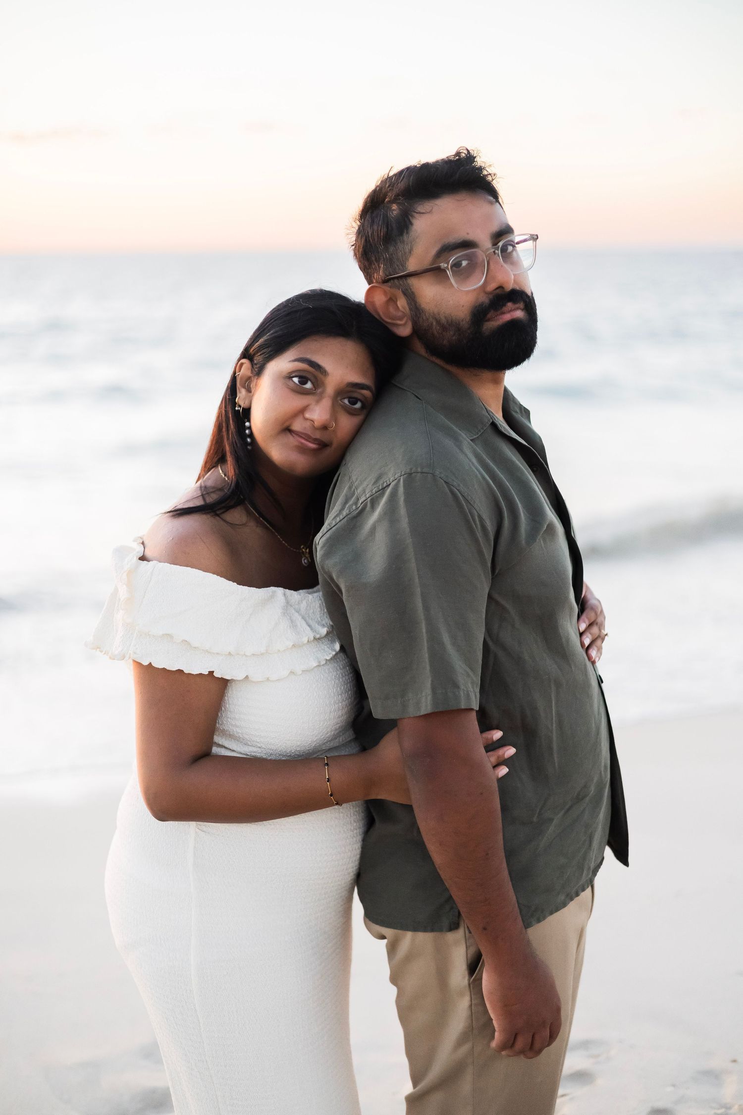 Indian couple posing back-to-back on the beach at dusk, woman in white off-shoulder dress leaning on mans shoulder.