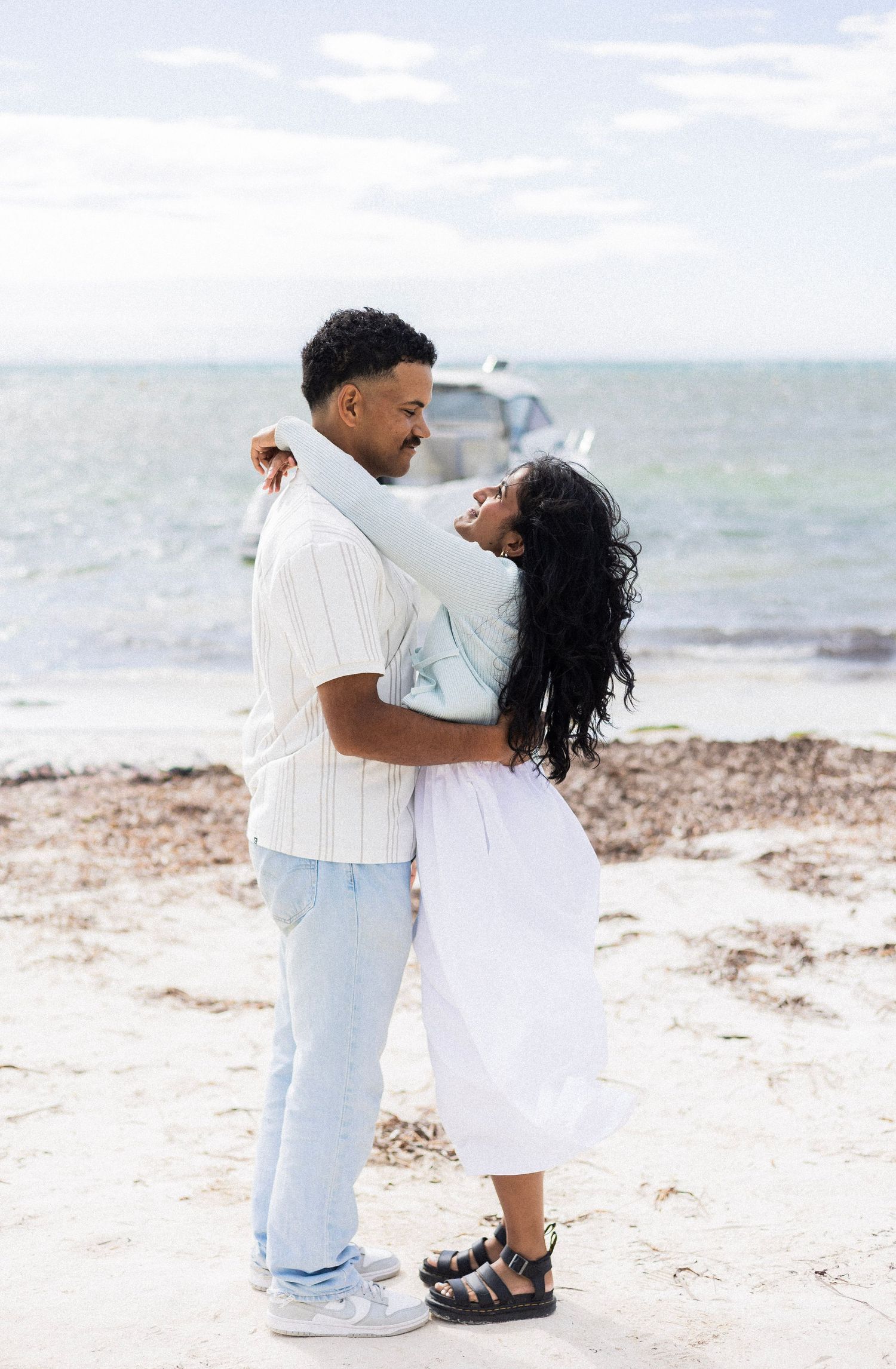 A couple embraces romantically on a sandy beach with ocean waves in the background.
