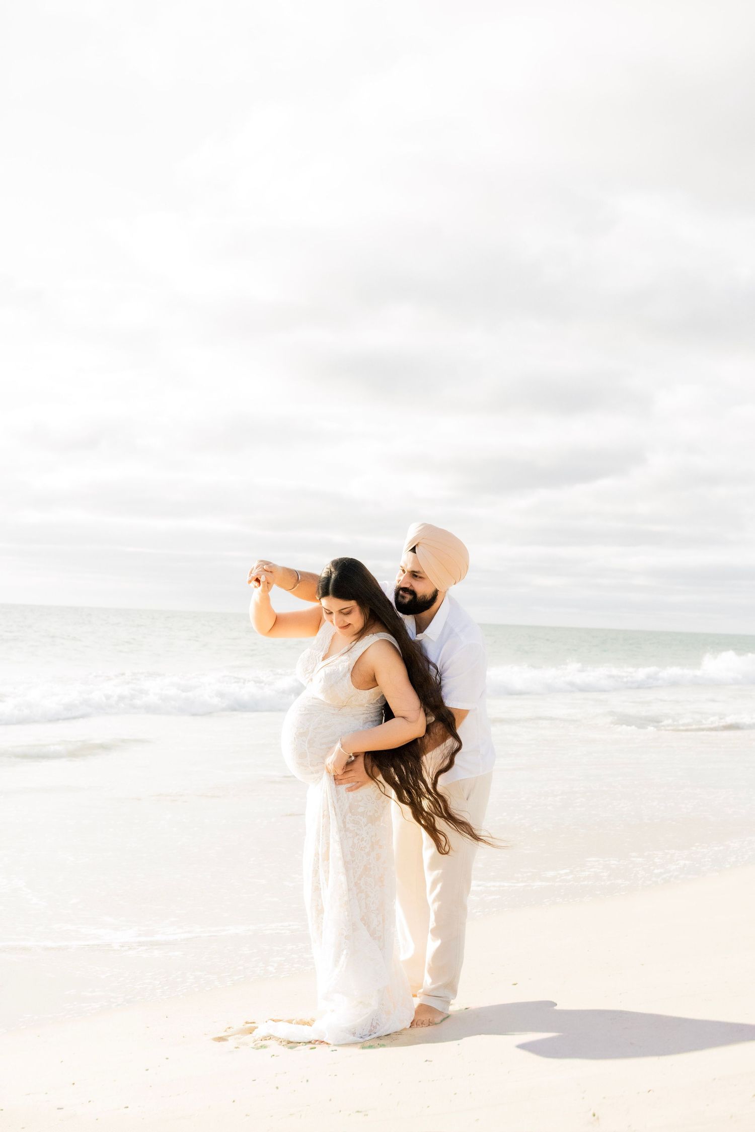 Partner in turban spinning pregnant woman in white lace dress on a beautiful sandy beach.