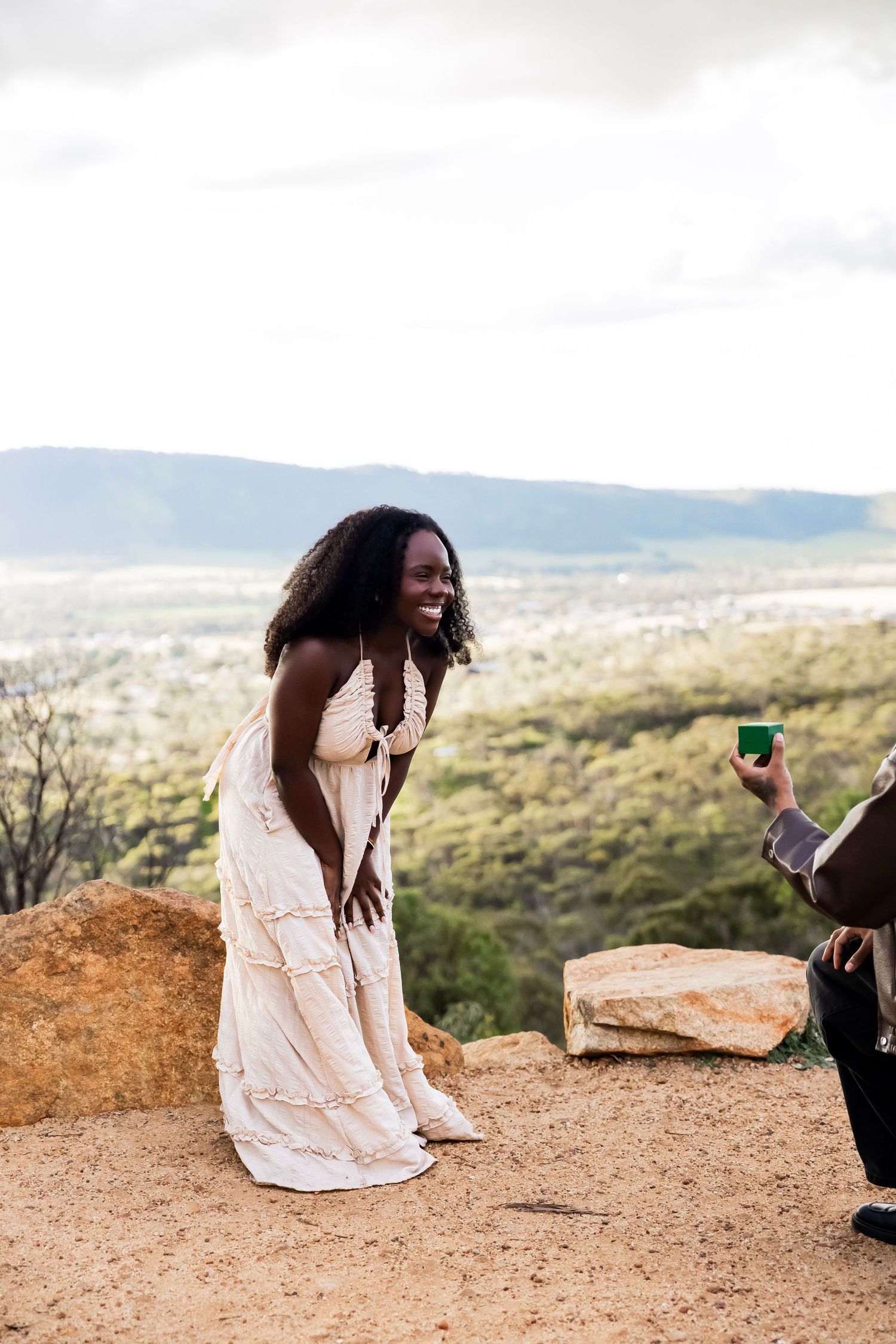 A man kneeling to propose to a smiling woman in a white dress on a scenic hilltop overlook.