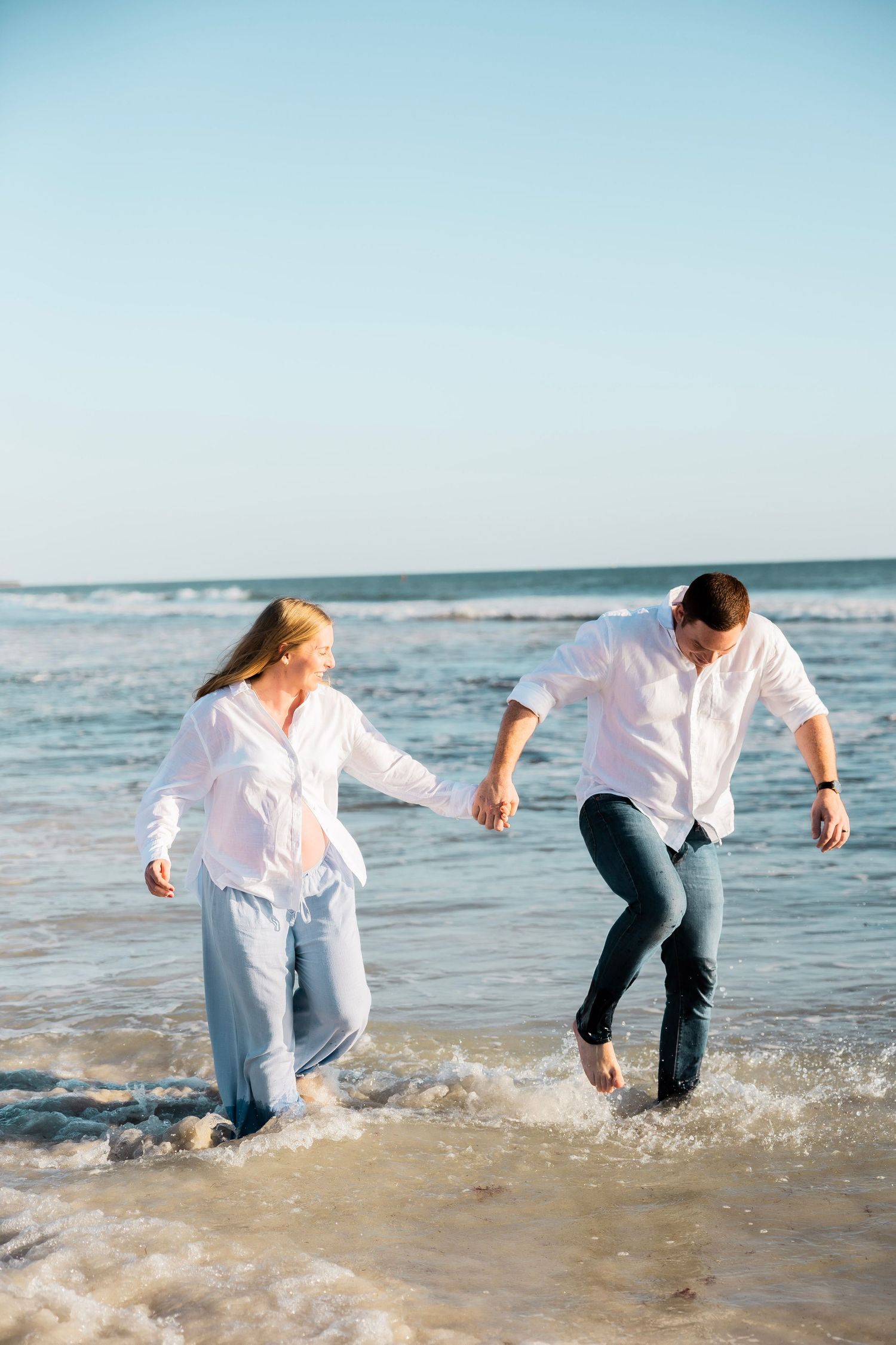 Laughing pregnant couple holding hands and splashing through ocean waves on a bright sunny beach.