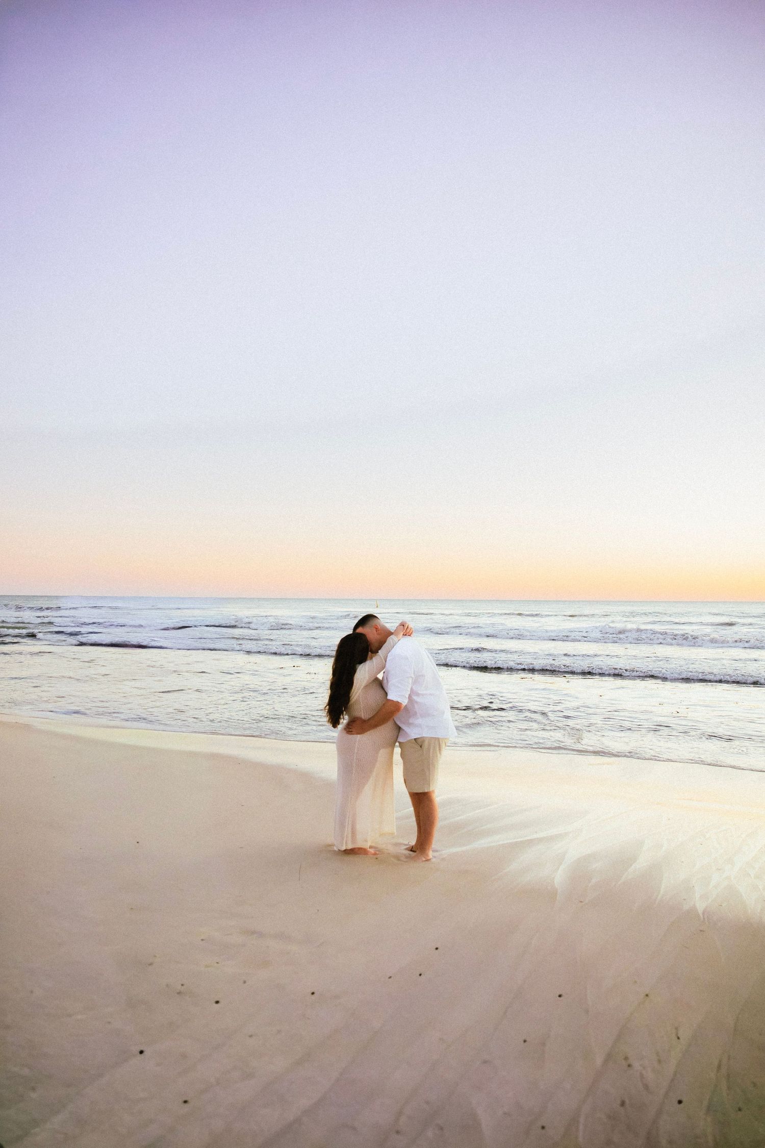 A couple embraces on a white sand beach at sunset during a maternity photoshoot.