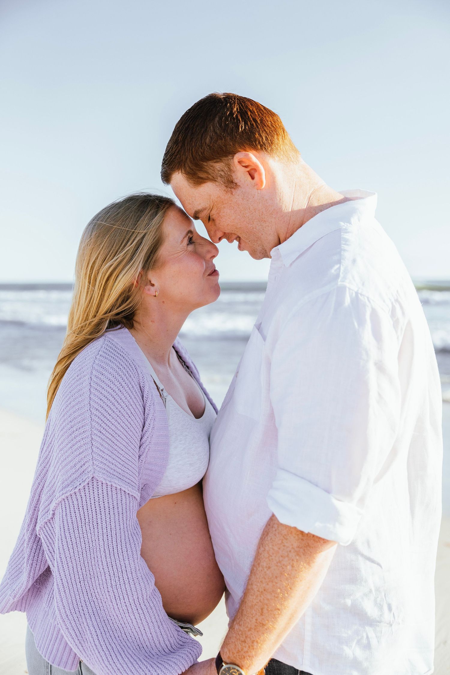 Pregnant couple touching foreheads romantically on a sunny beach with ocean waves in the background.
