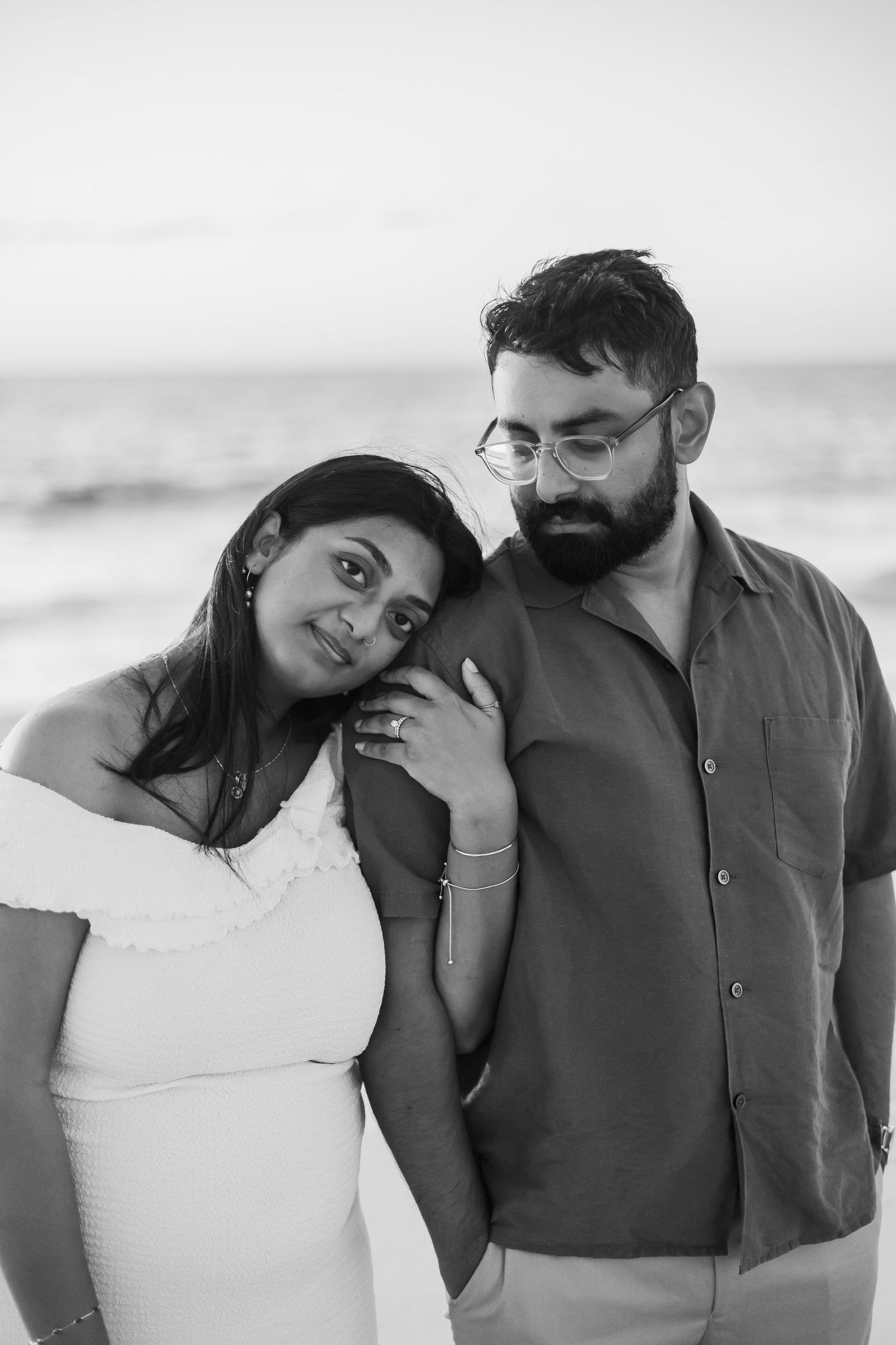 Black and white beach portrait of a couple, woman in white dress leaning on bearded man wearing glasses.