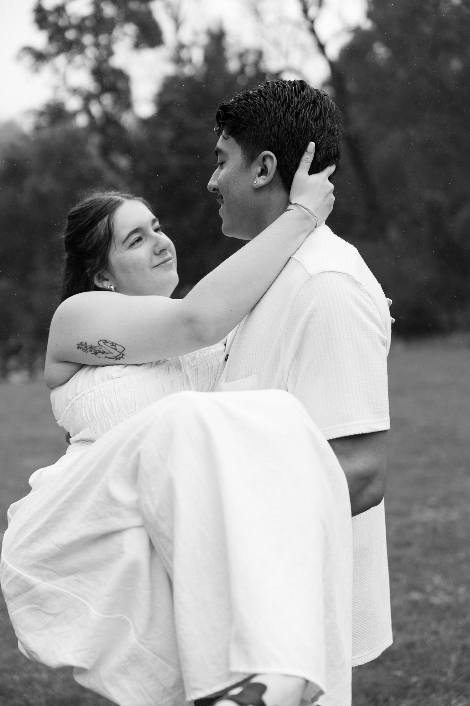 A man carries a woman in a white dress in his arms while she tenderly holds his face in a romantic black and white photo.