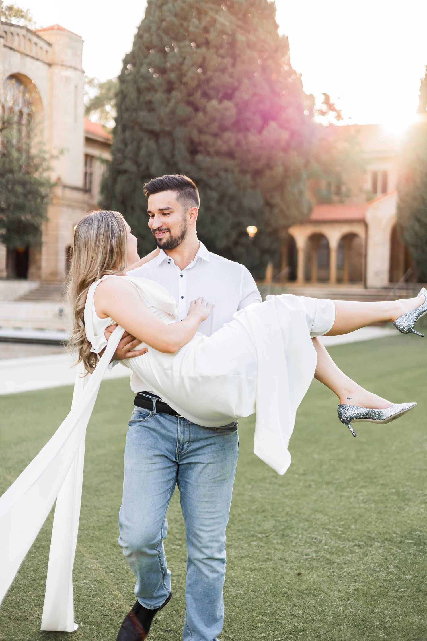 Man carrying smiling woman in white dress on a green lawn with warm golden sunlight behind them.