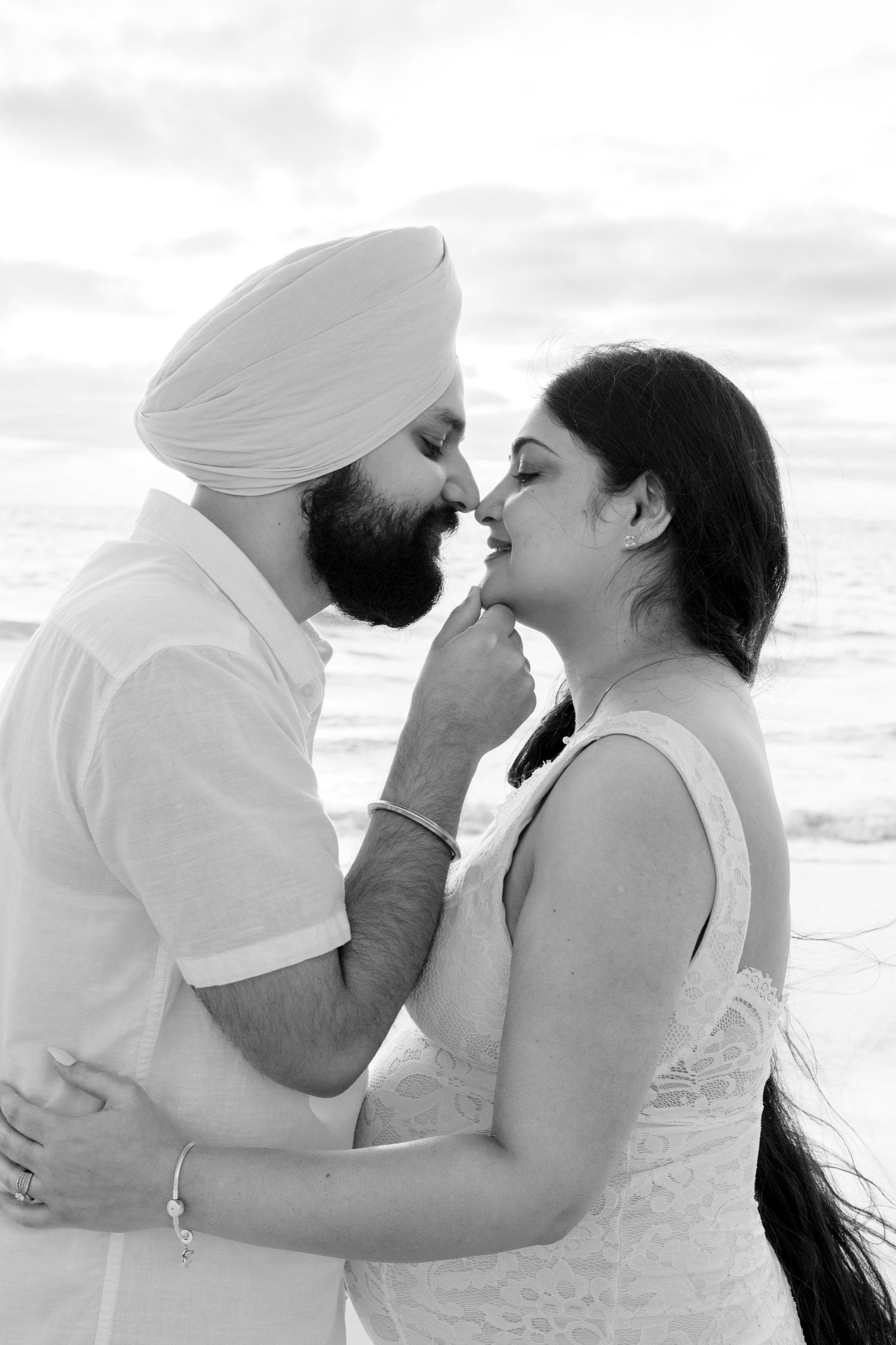 Couple sharing intimate moment on beach, man in turban tenderly touching womans lips, black and white.