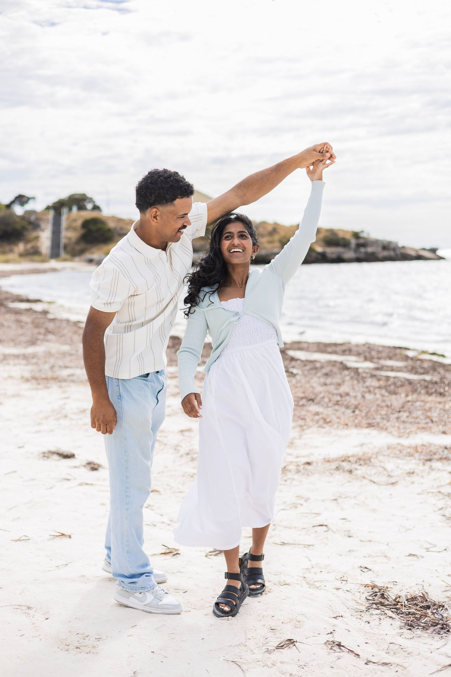 A couple dances joyfully on a sandy beach as the man twirls the woman with the ocean behind them.