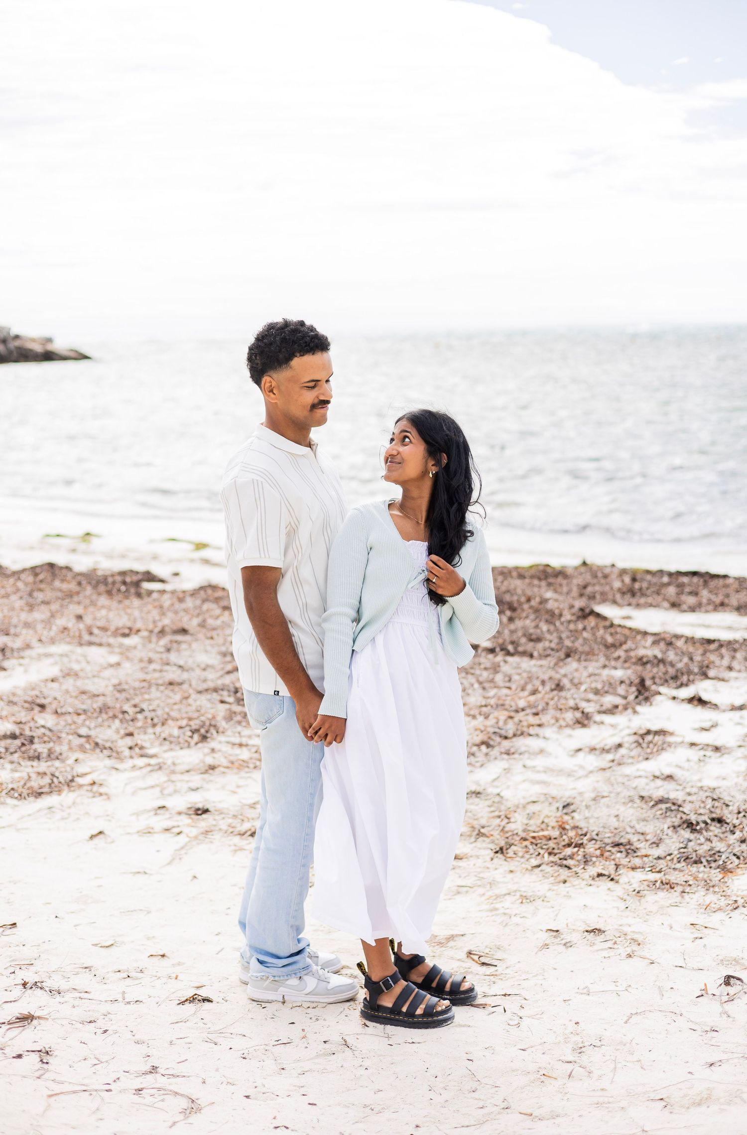 A couple holds hands and gazes at each other lovingly on a serene sandy beach with calm ocean waters.