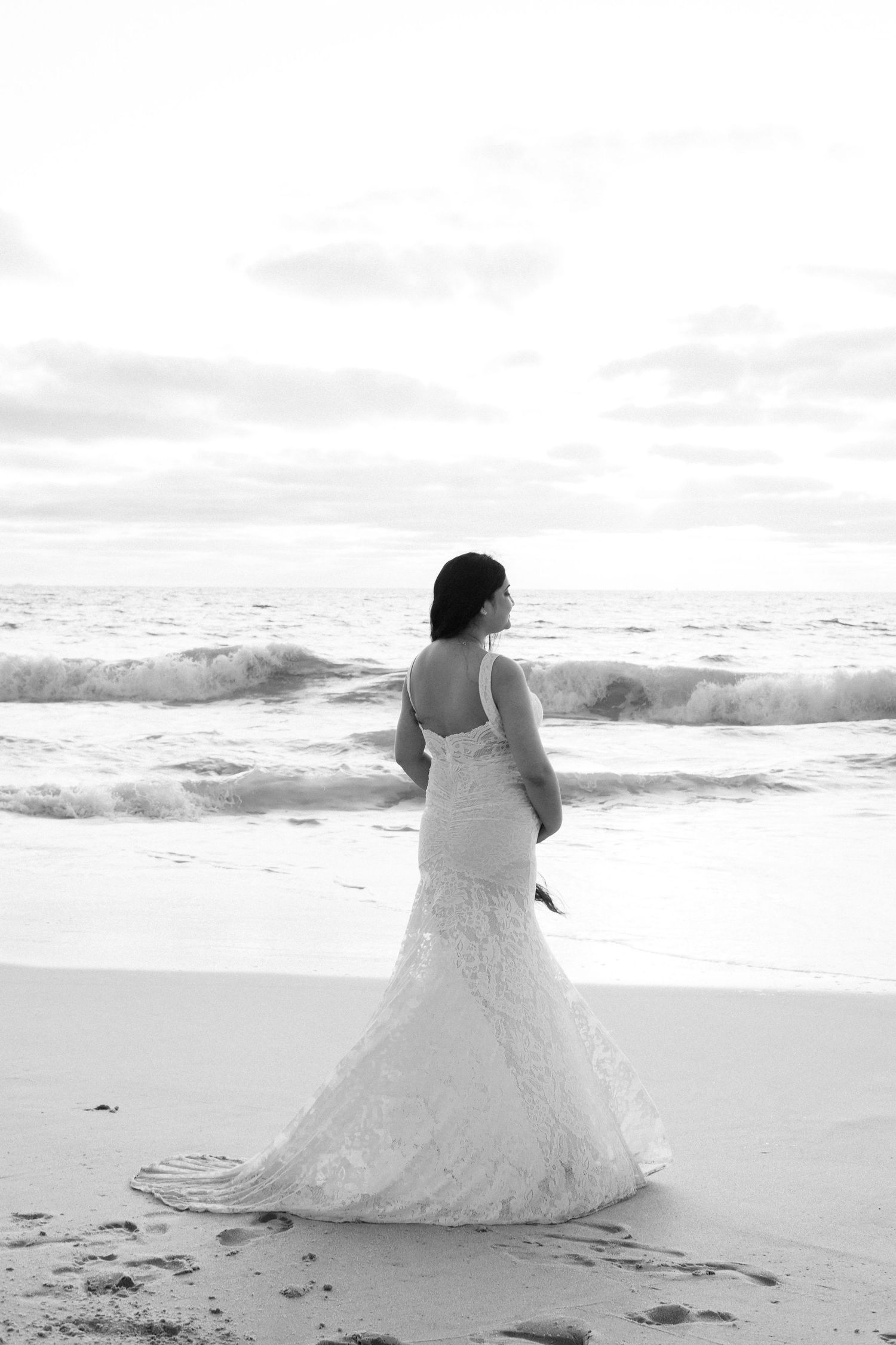 Bride in white lace mermaid gown standing on beach shore gazing at ocean waves, black and white photo.