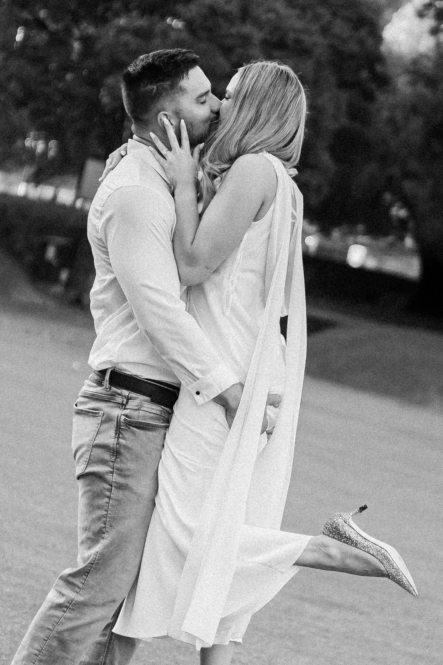 Black and white photo of a couple sharing a passionate kiss on a green lawn outdoors.