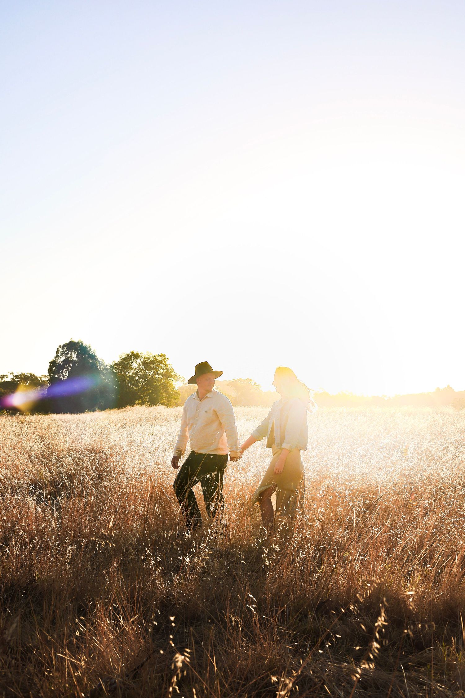 Couple walking hand in hand through sun-drenched golden grass field at sunset with lens flare.