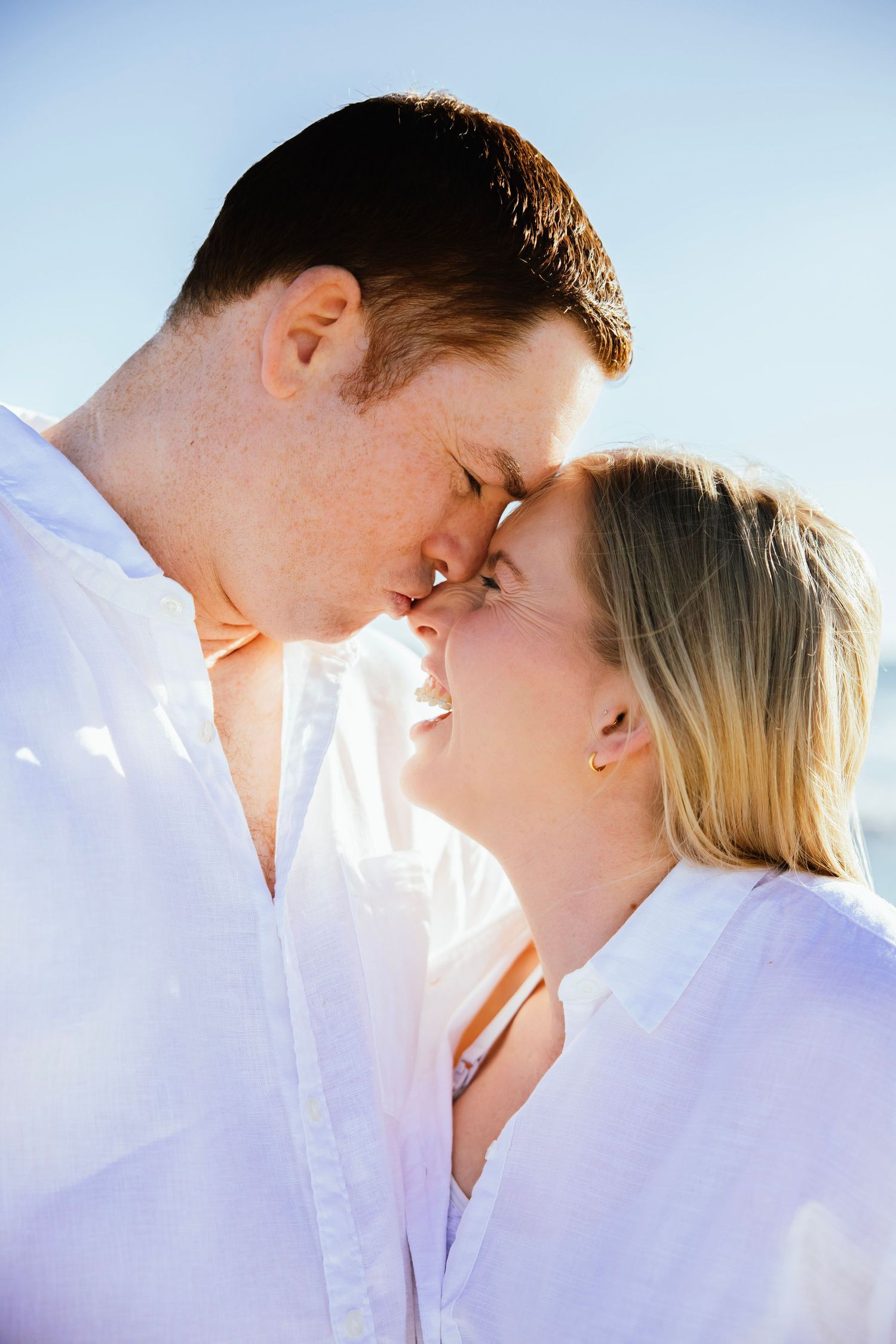 Romantic couple in white shirts leaning in for a kiss on a bright sunny beach.