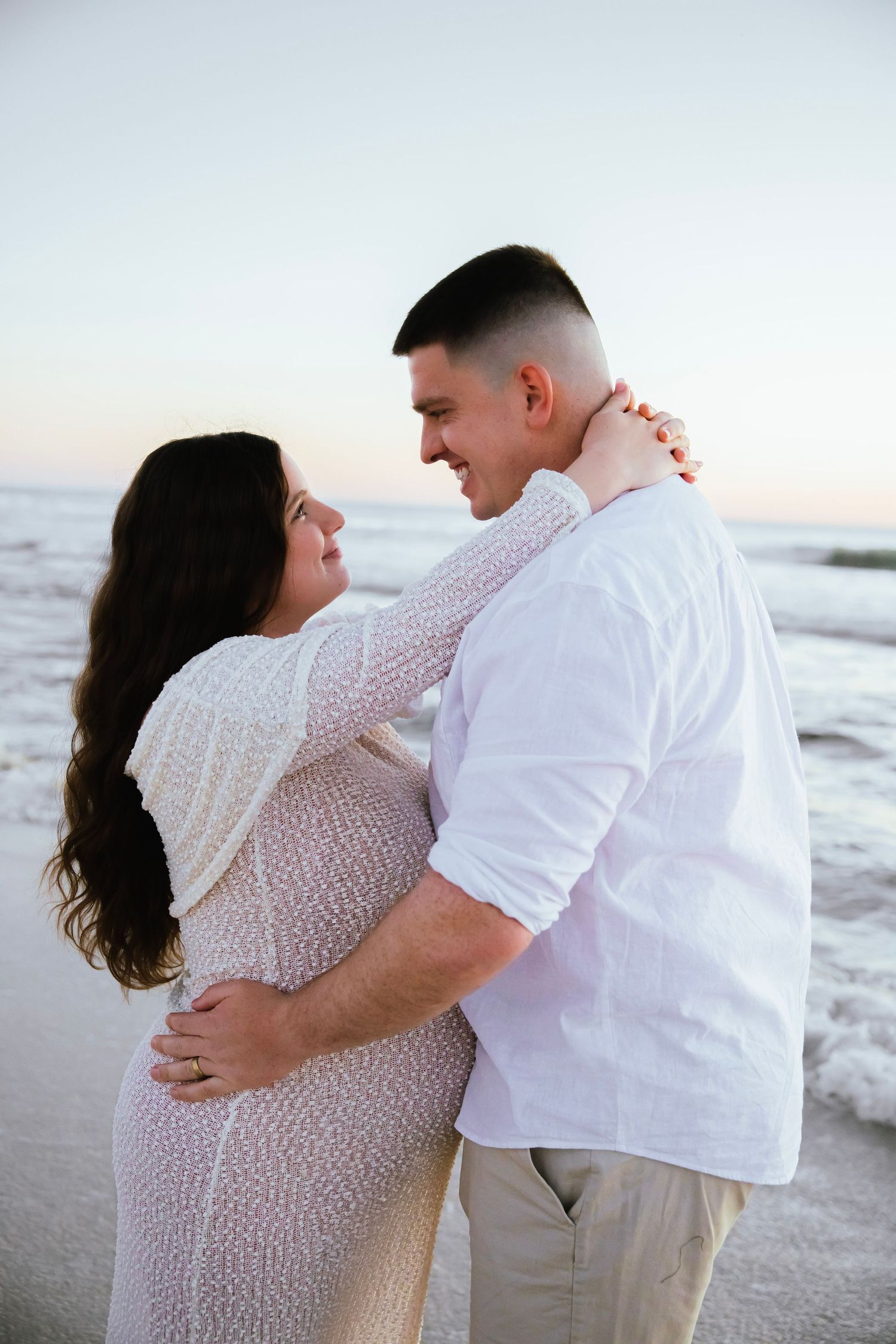 Pregnant couple embracing and smiling at each other on beach at dusk, dressed in white outfits.