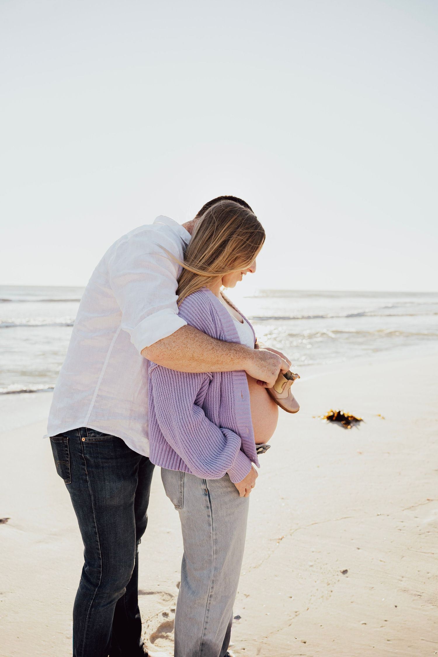 Couple embracing on a sandy beach, man hugging pregnant woman from behind near ocean waves.