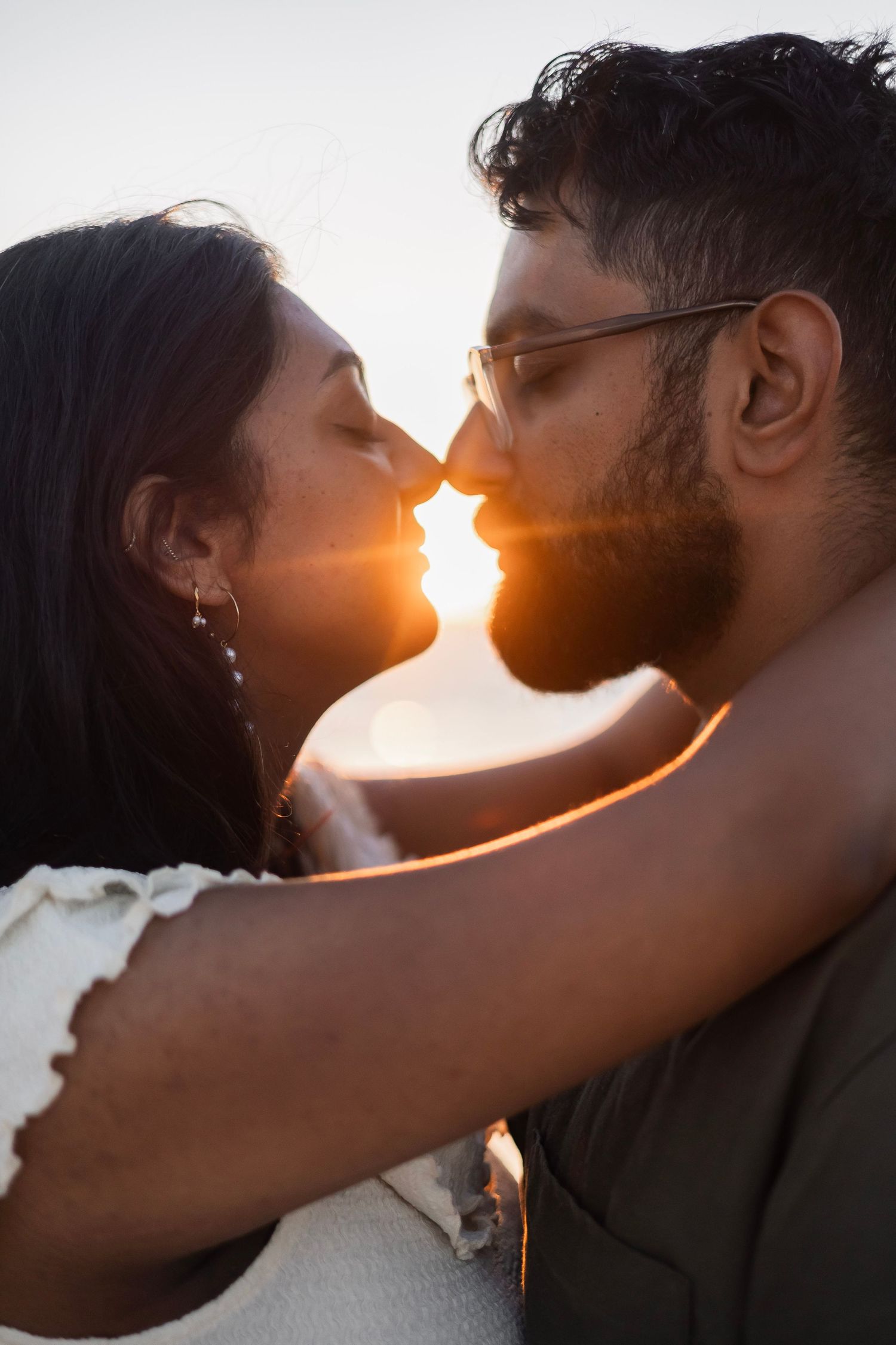 Couple sharing a romantic sunset kiss, backlit by golden light, man wearing sunglasses embracing woman outdoors.