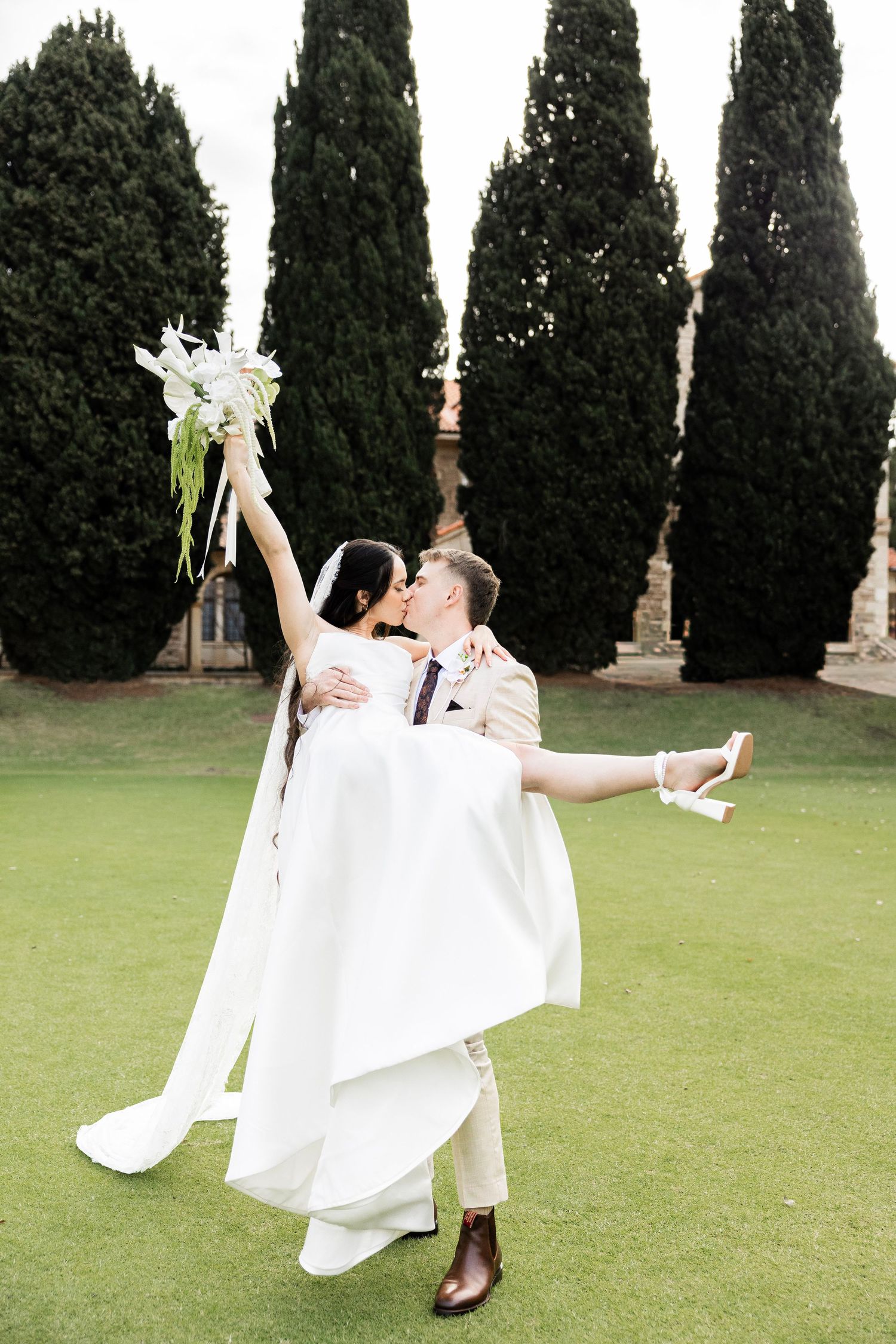 Groom lifts bride in white gown as they kiss on green lawn with tall cypress trees backdrop.