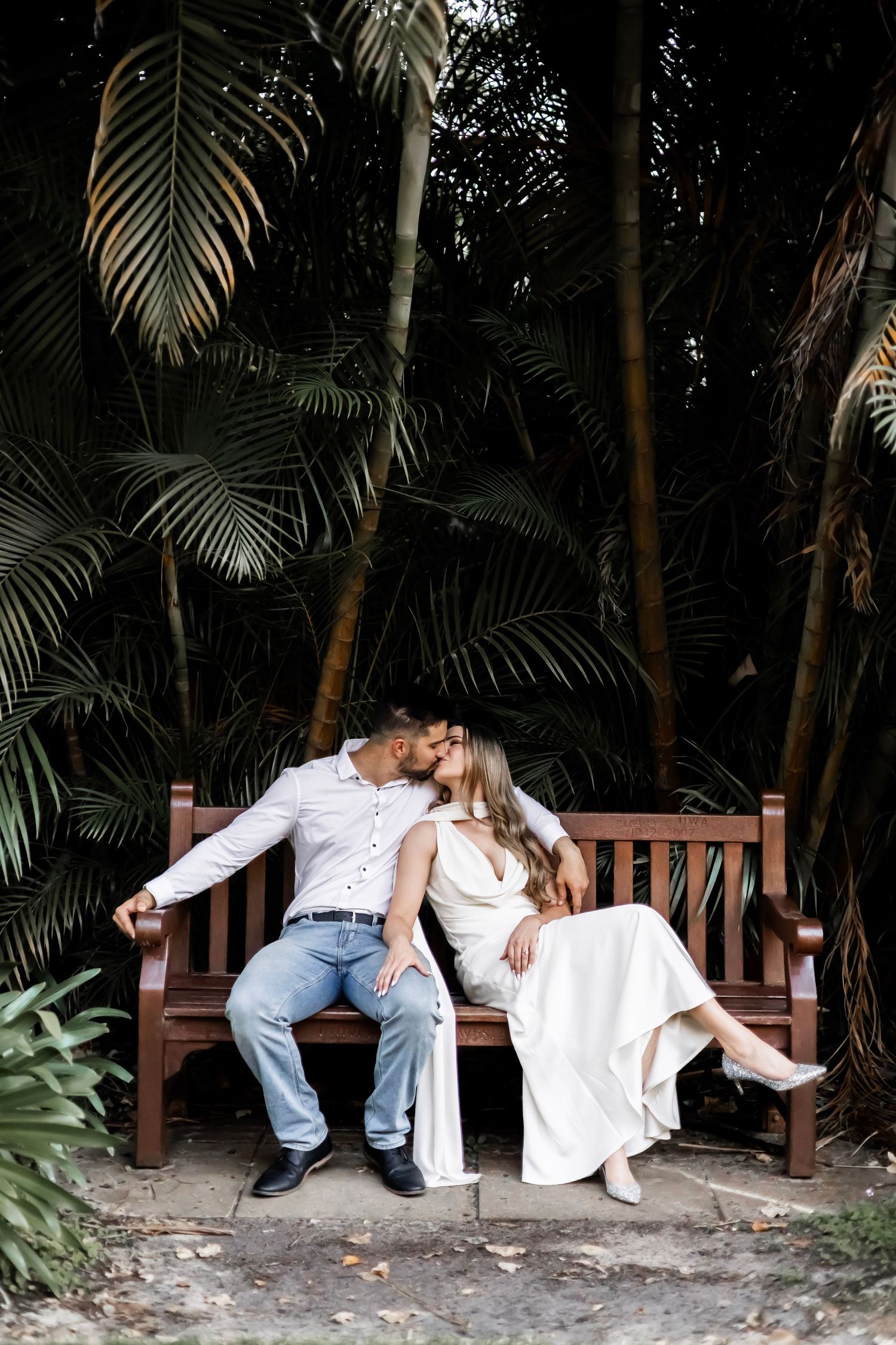 Couple sharing a romantic kiss on a wooden bench surrounded by lush tropical palm trees.