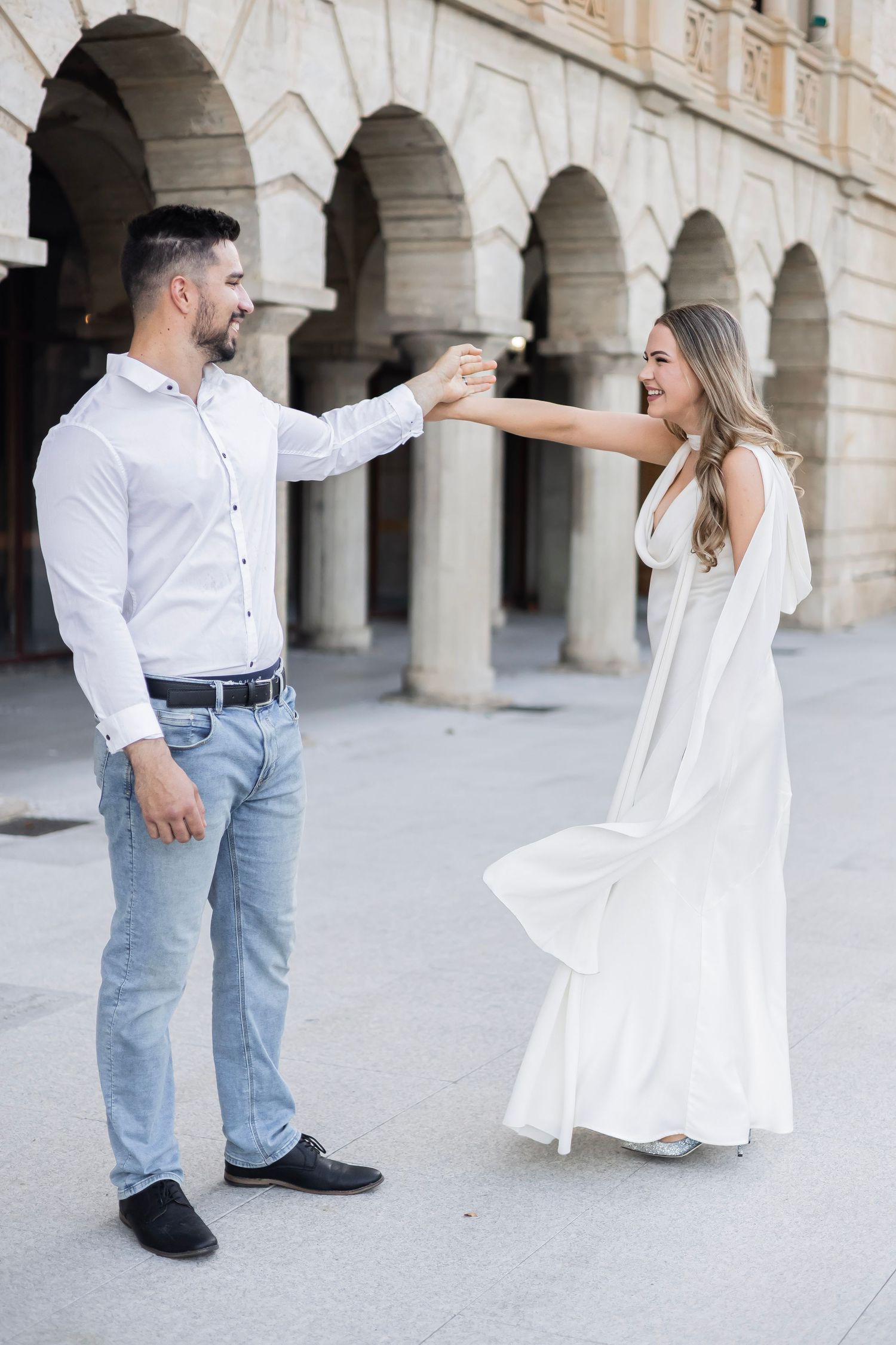 Joyful couple dancing and holding hands in front of grand stone arched building facade.